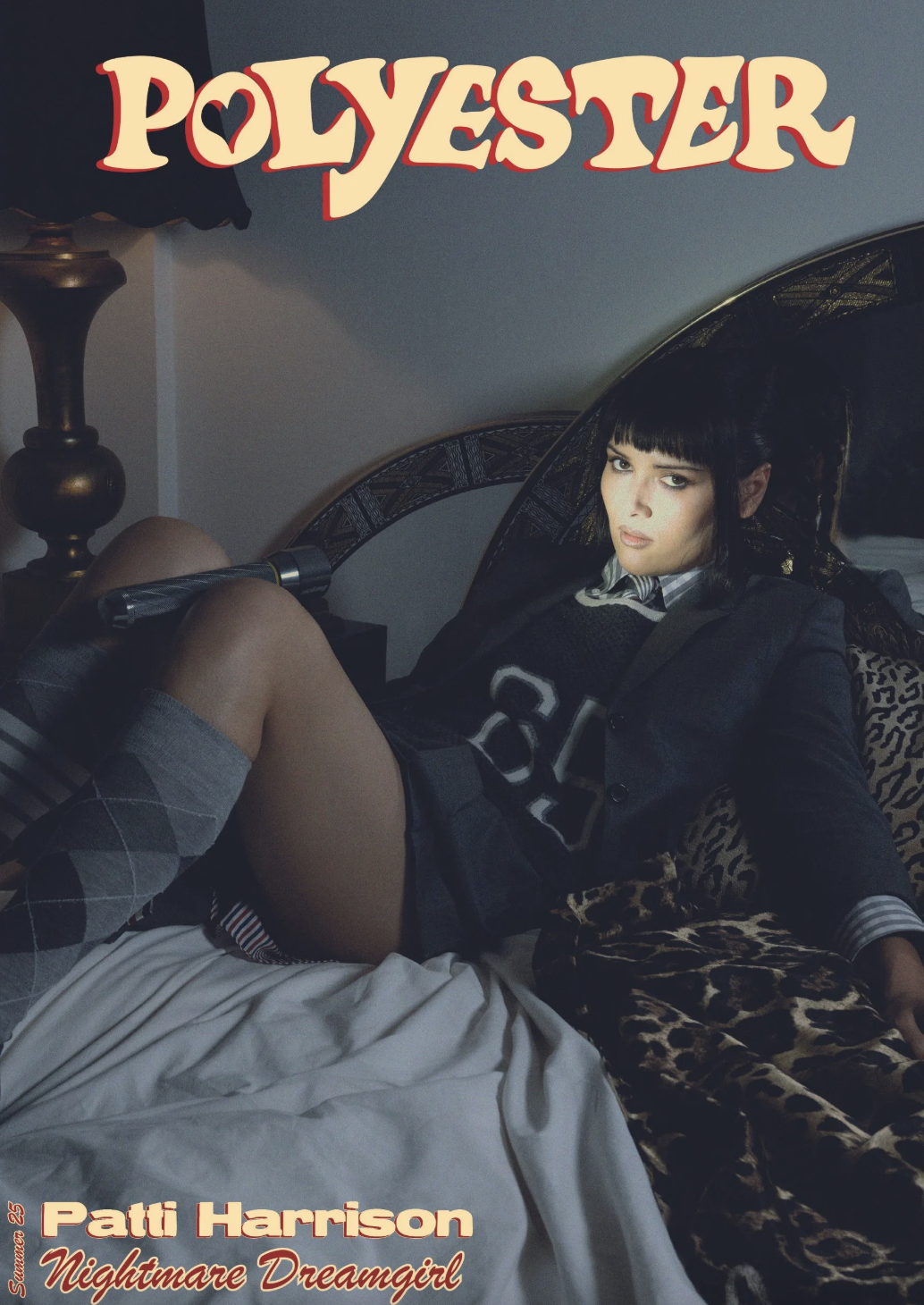 A young woman with black hair and bangs is sitting on a bed, wearing a black blazer, a sweater with a number on it, short shorts, and argyle socks. She is looking at the camera with a serious expression, and the bed has leopard-print blankets. There is a mirror behind her and a lamp on a nightstand next to the bed. The image has text that reads "Polyester," "Patti Harrison," and "Nightmare Dreamgirl."