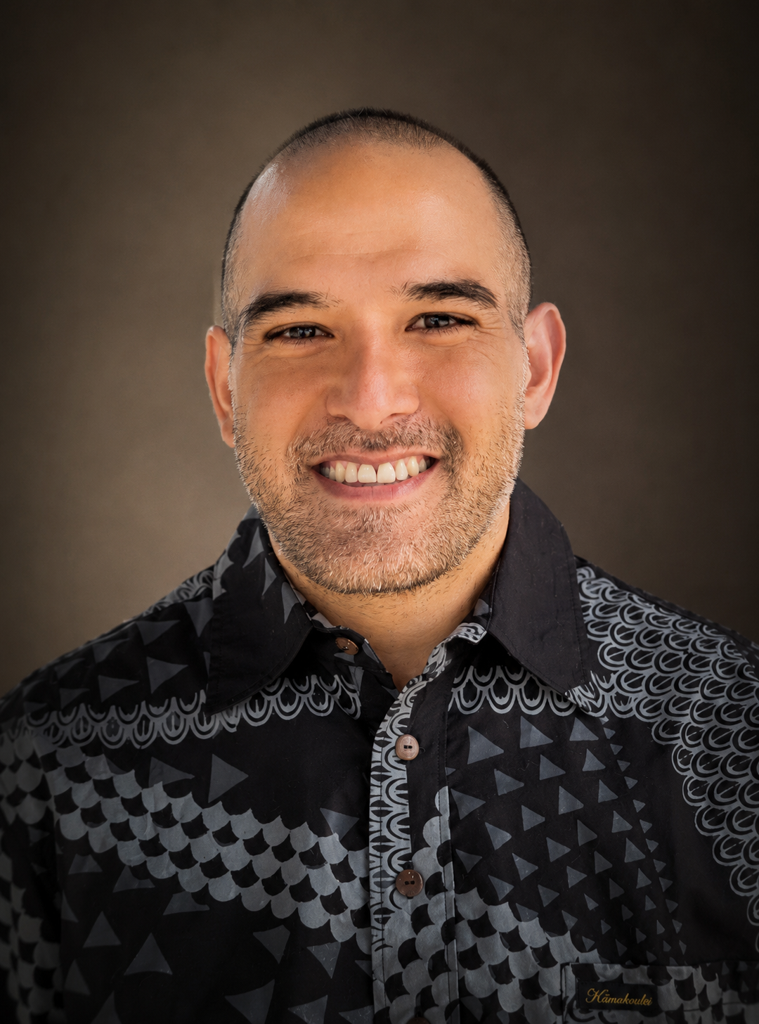 Close-up portrait of a smiling man with a beard and short hair, wearing a black patterned shirt, against a dark background.