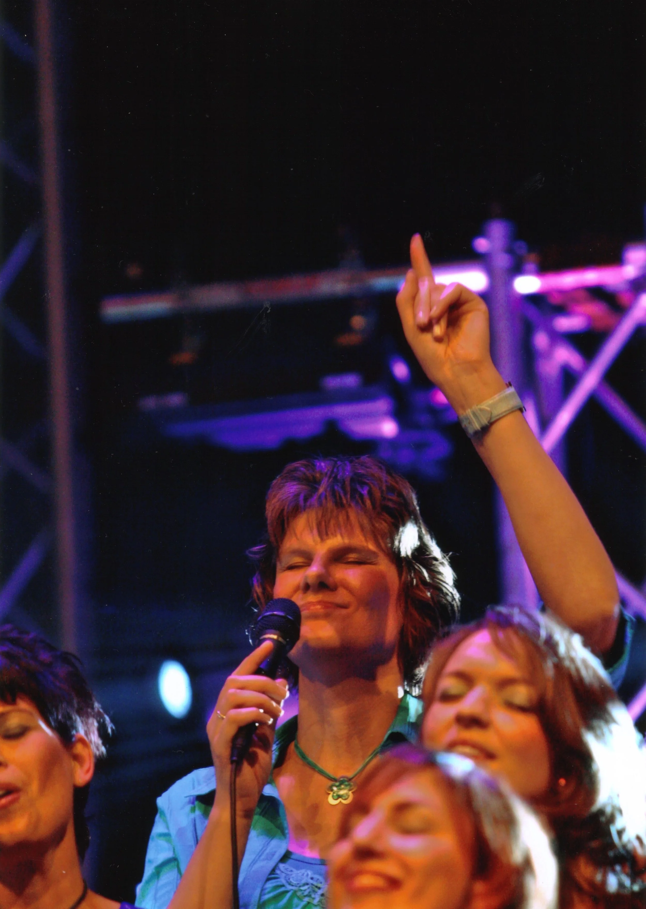 Een vrouw zingt in een koor. Ze heeft haar ogen dicht, houdt een microfoon vast en wijst met een hand omhoog.