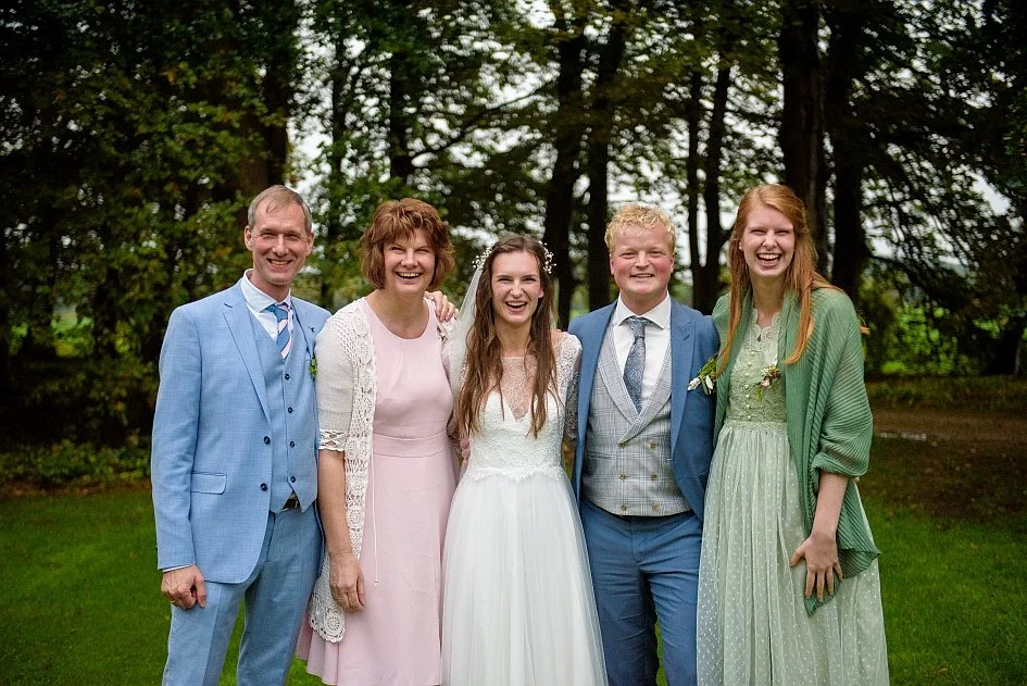 Vijf personen staan naast elkaar en lachen naar de camera. De middelste vrouw heeft een trouwjurk aan. De anderen hebben formele kleding aan.