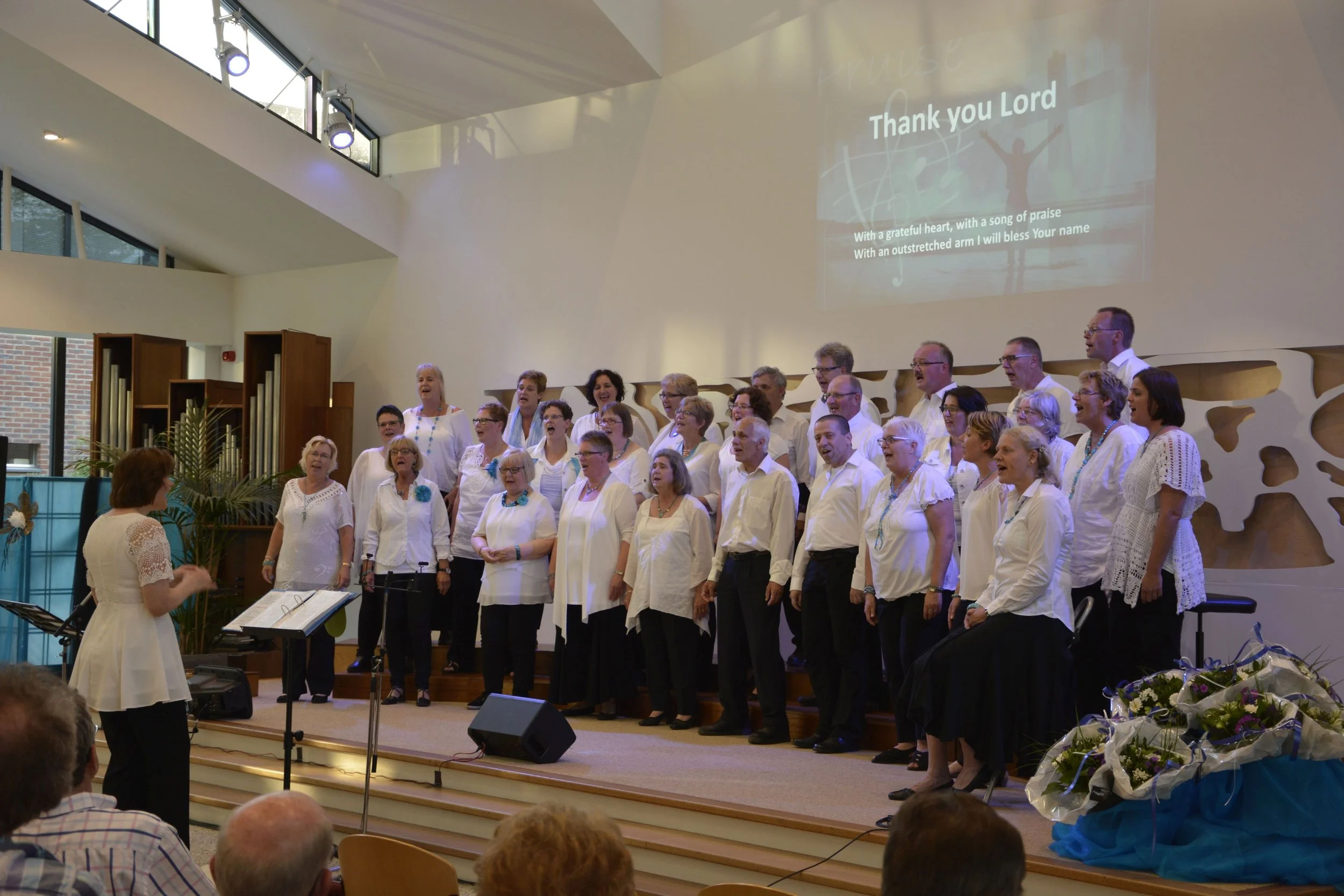 Een groot koor staat op een podium te zingen. Op de beamer staan de woorden ' thank you lord' .