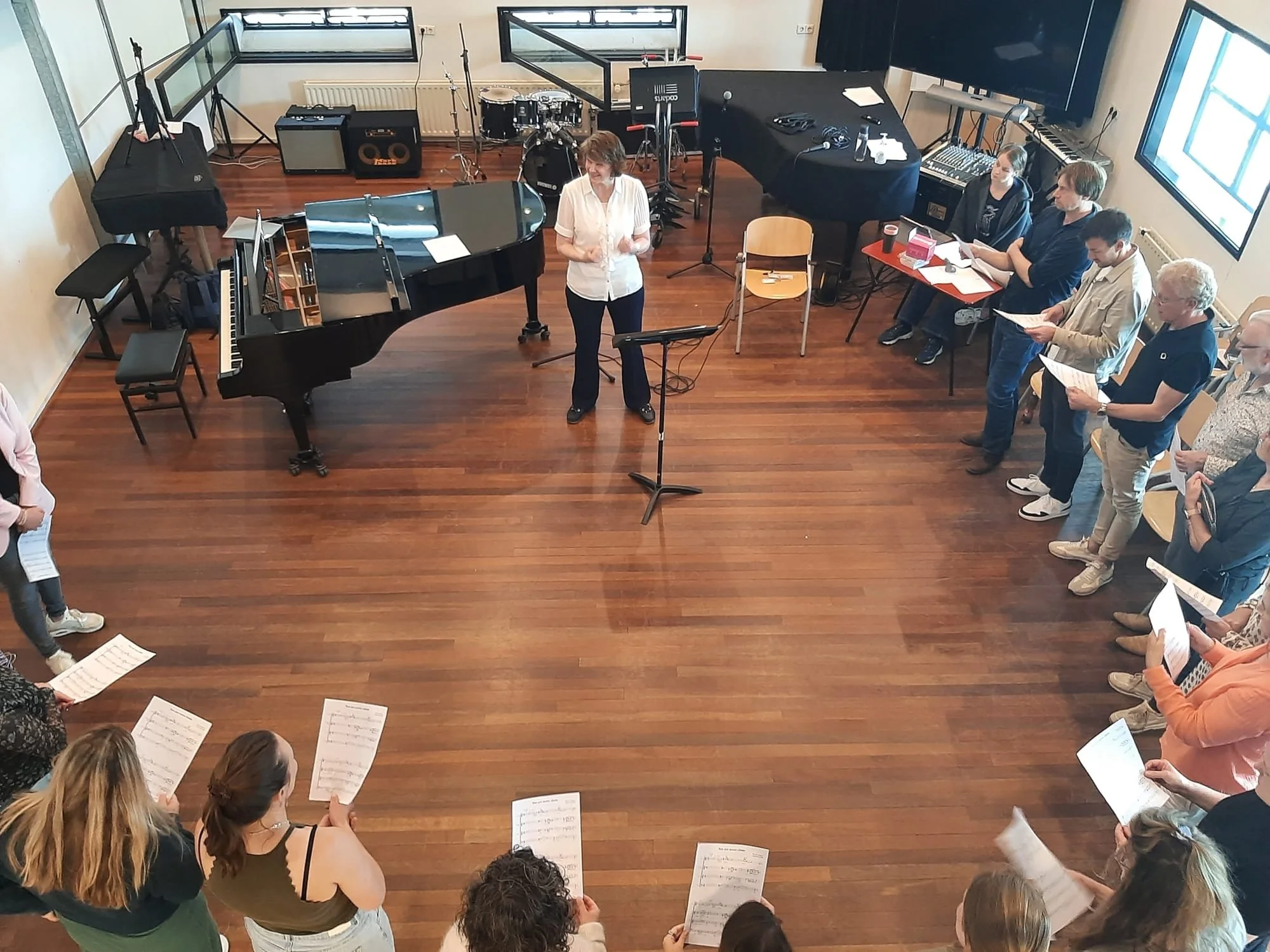 Een foto van boven genomen. Een groep mensen staan in een kring met bladmuziek in hun handen. Een dirigent leidt.