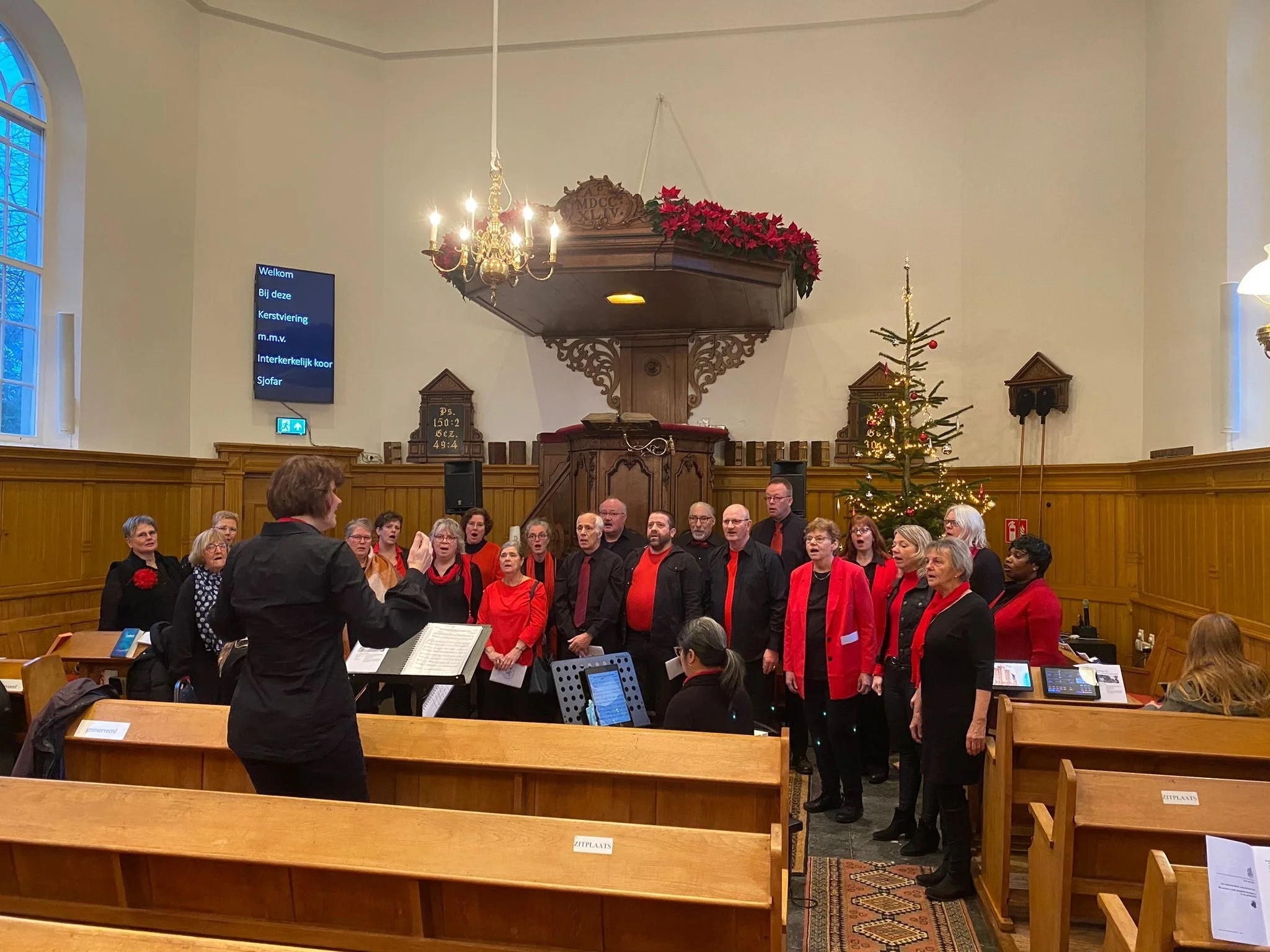 Een groot koor, gekleed in zwart en rode kleding, zingt samen in een kerk.