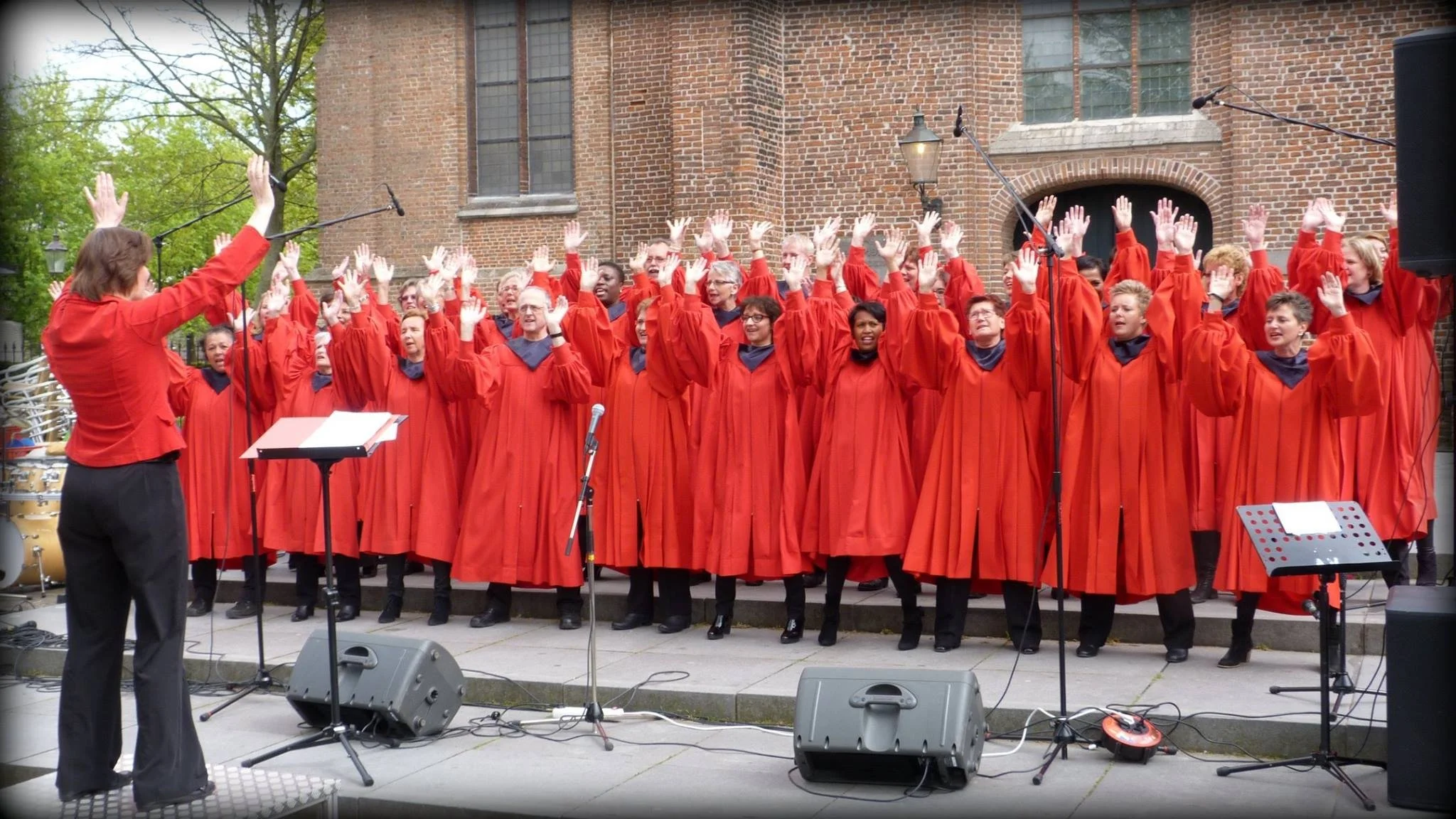 Een groot koor gekleed in rode robes zingt samen. Hun handen zijn in de lucht.