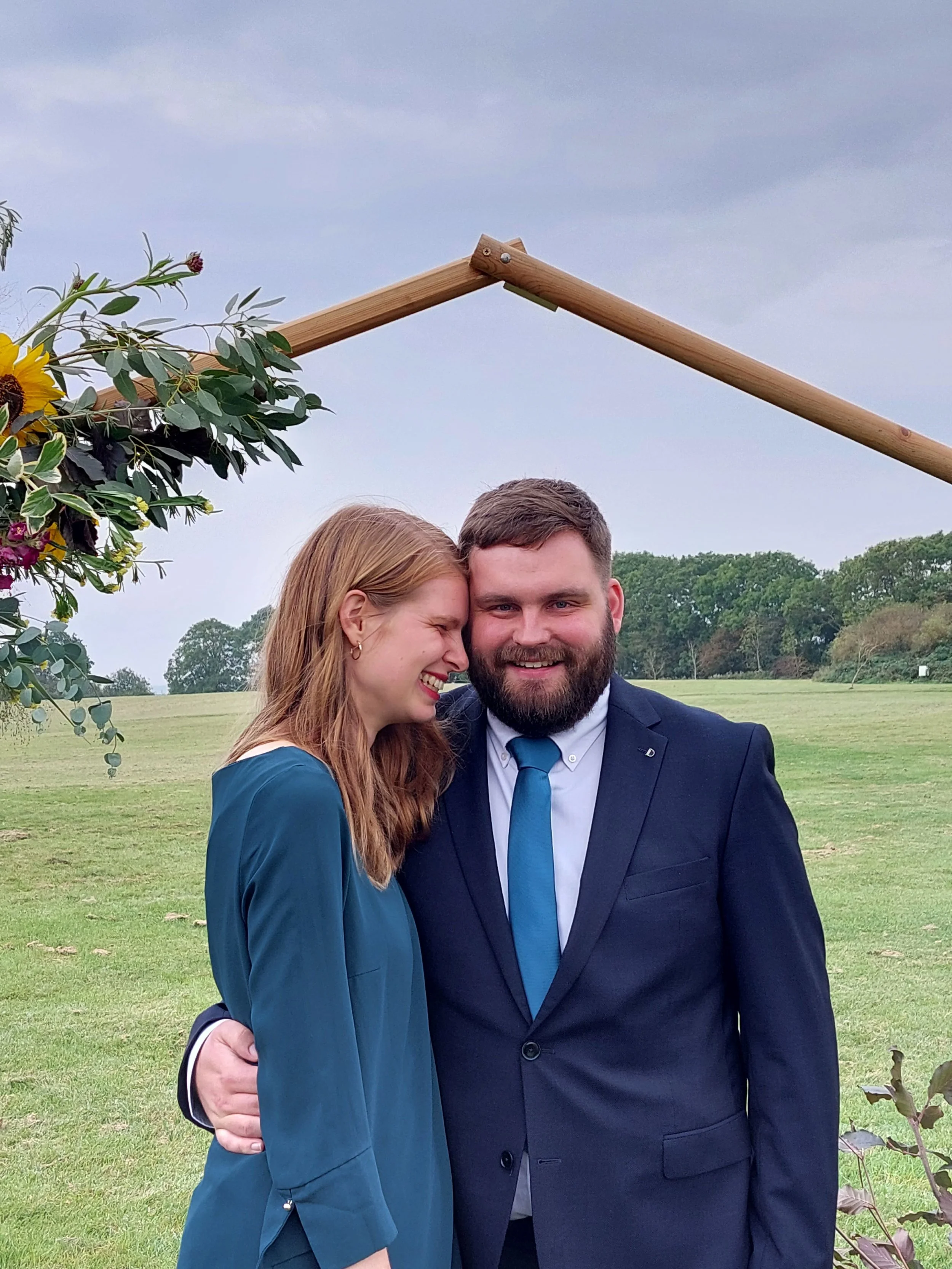 Een stel staat op de foto. De man kijkt naar de camera. De vrouw leunt haar hoofd tegen zijn hoofd.