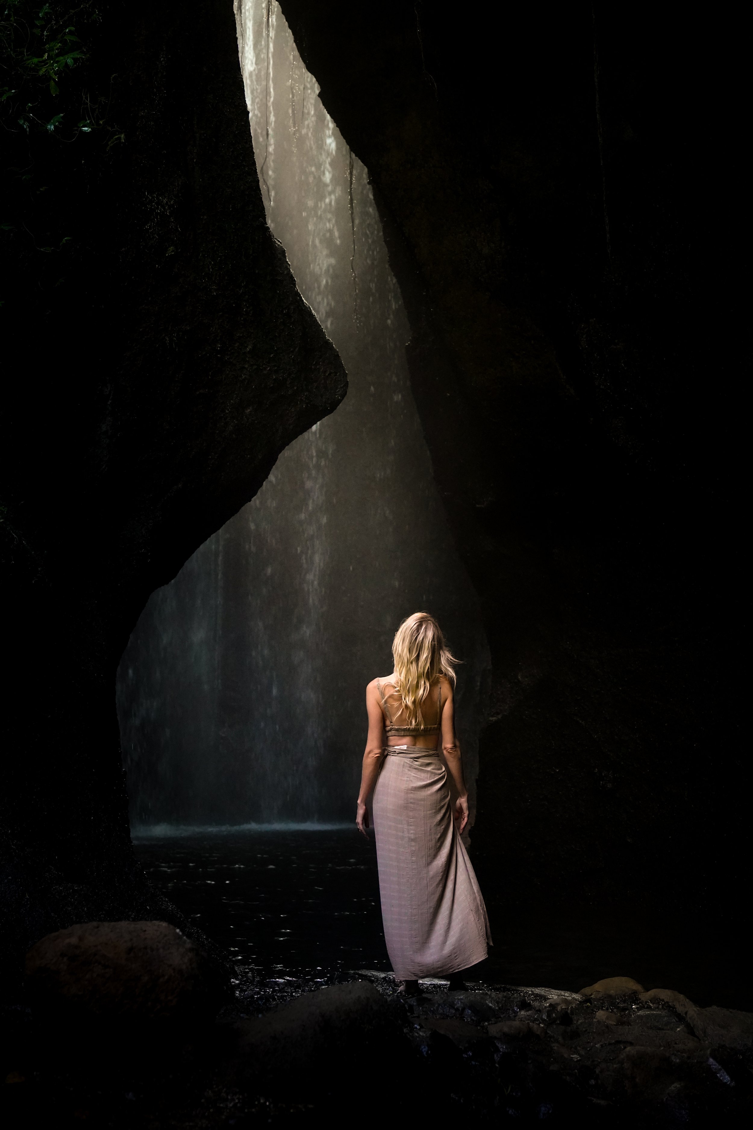 A woman with long blonde hair wearing a beige skirt and top standing in front of a large waterfall inside a dark cave.