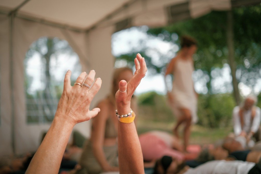 Close-up of hands with rings and bracelets raised during a yoga class, with people practicing yoga on mats outdoors under a canopy.