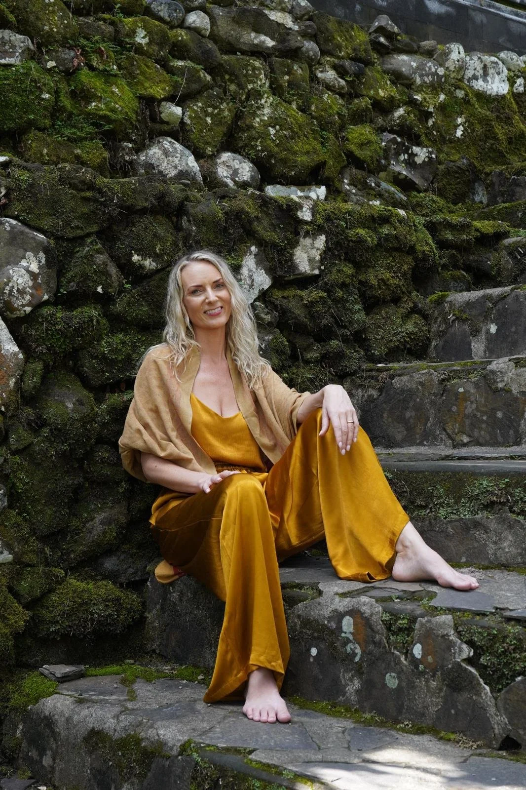 A woman with blonde curly hair wearing a mustard yellow dress and matching pants, sitting barefoot on stone steps against a moss-covered stone wall, smiling at the camera.