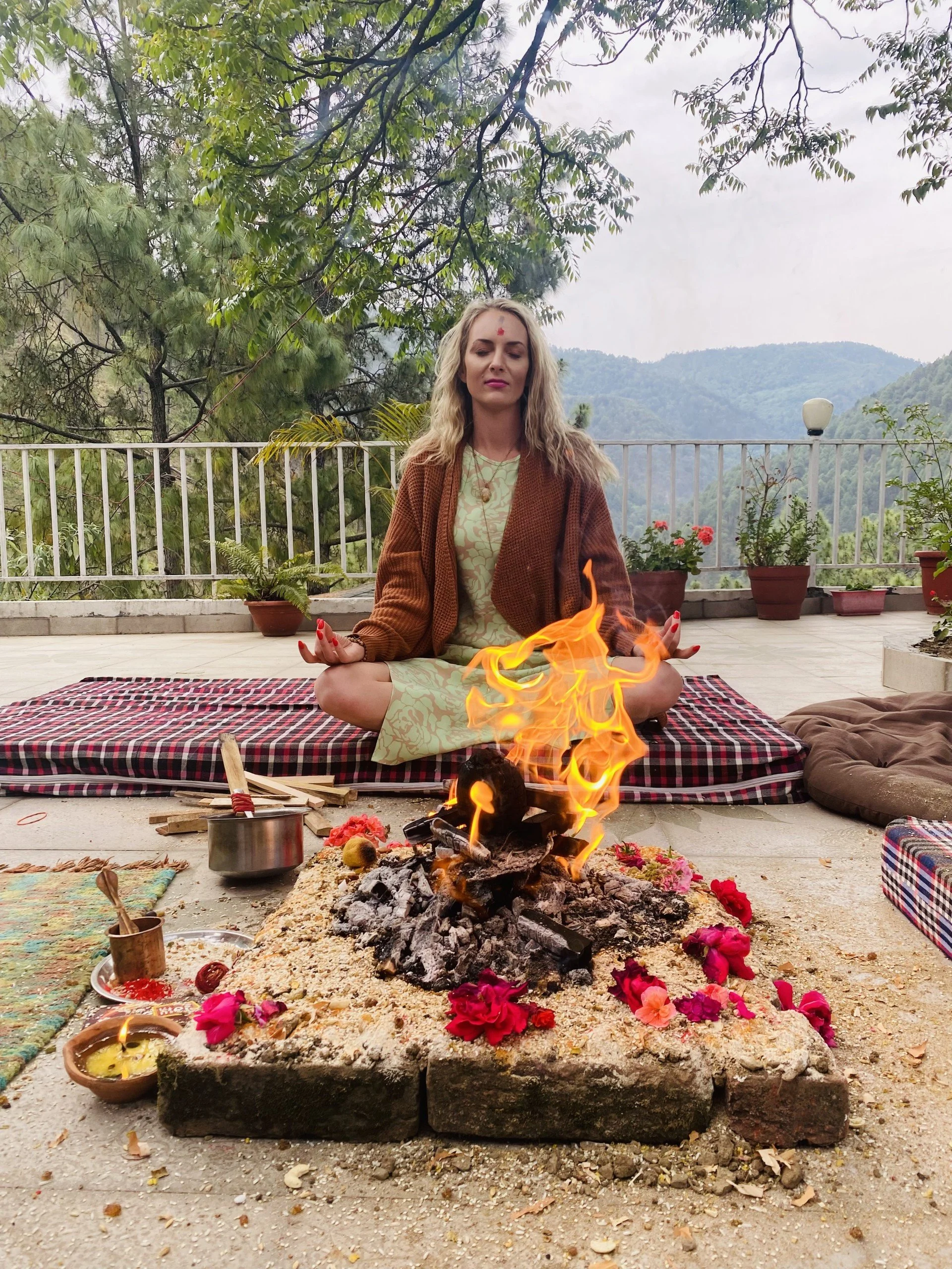 A woman practicing meditation outdoors next to a fire, with potted plants and mountains in the background.