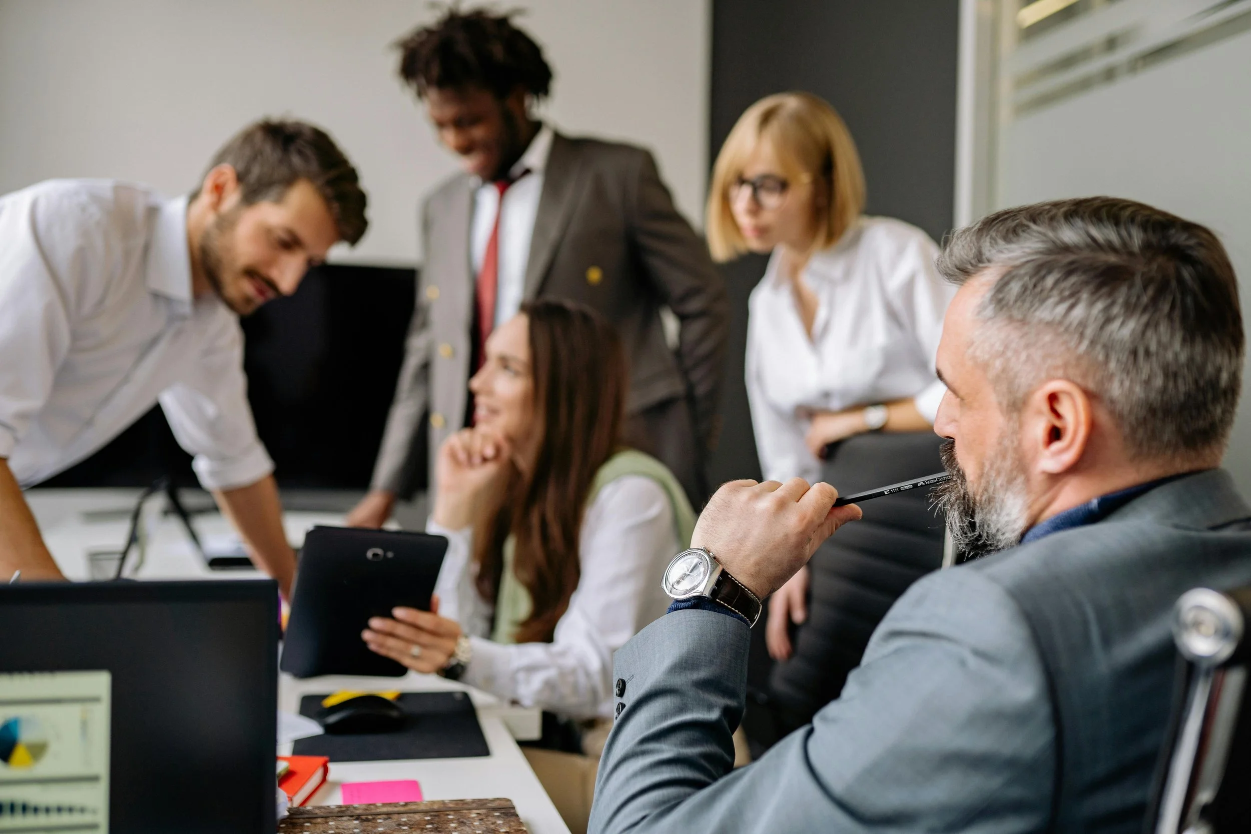 A diverse group of five professionals gathered around a desk in an office, engaging in a collaborative discussion. Some are standing, others are seated, focusing on a tablet and documents.