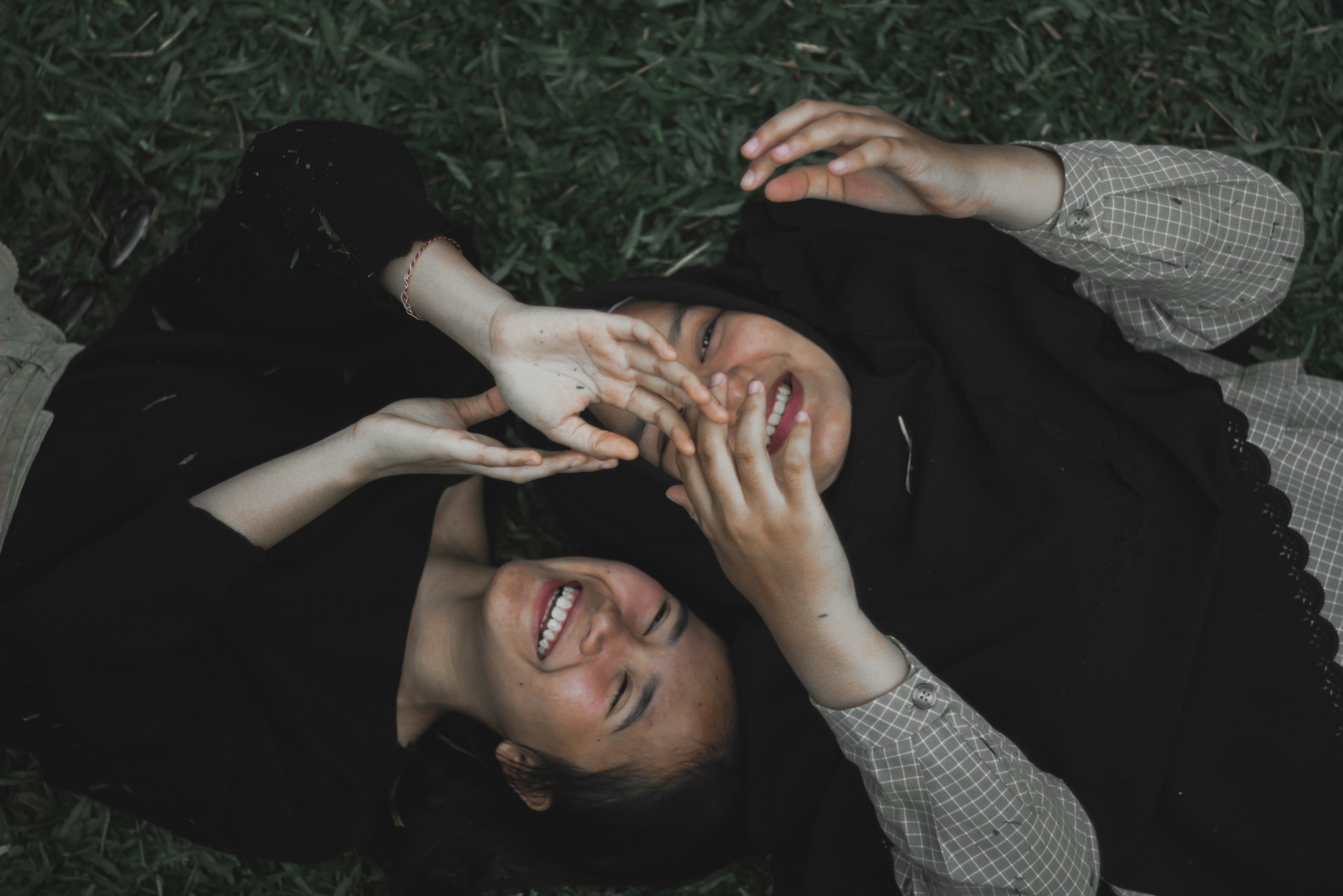 Two young women lying on grass, smiling and making peace signs with their hands, enjoying a moment outdoors.