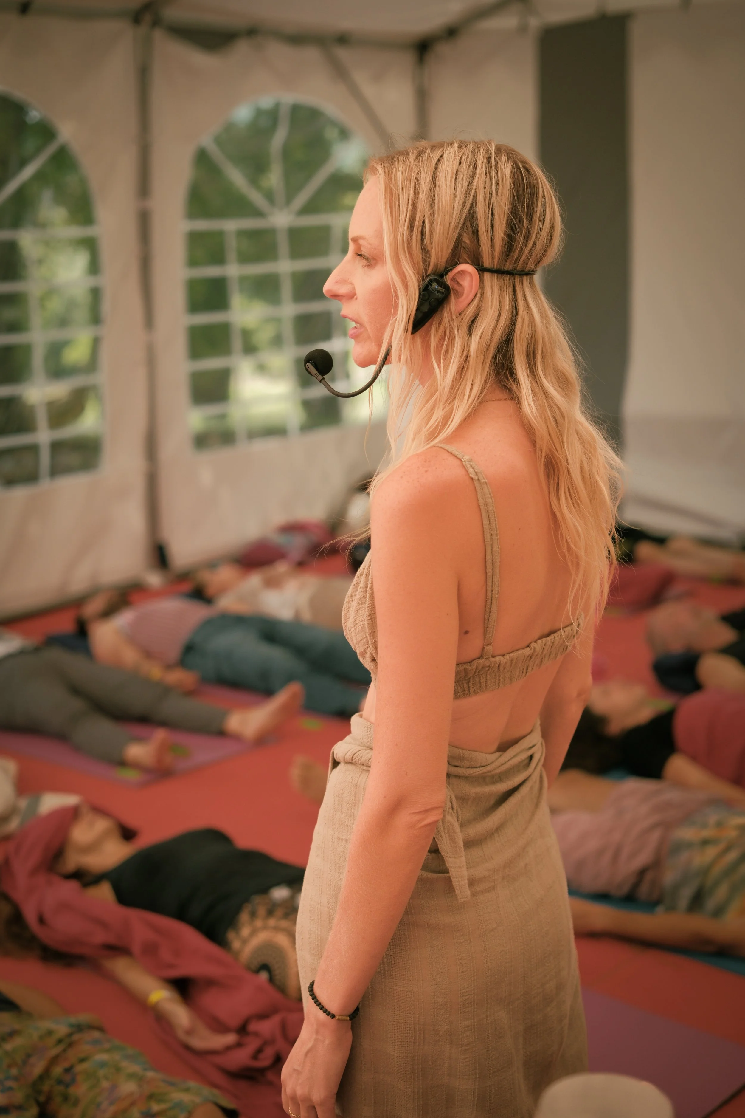 A woman with long blonde hair and a headset speaks in front of a group of people meditating inside a tent with large windows.