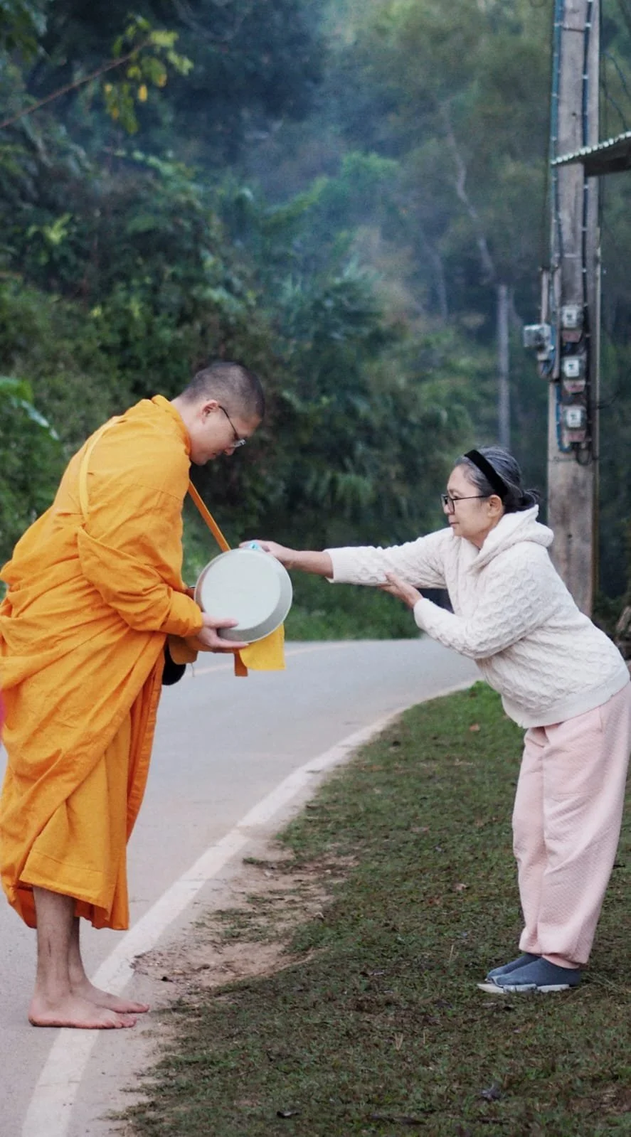 A woman wearing glasses and a white sweater is bowing with her hands together, receiving an alms bowl from a Buddhist monk dressed in an orange robe, standing barefoot on a roadside with green trees in the background.