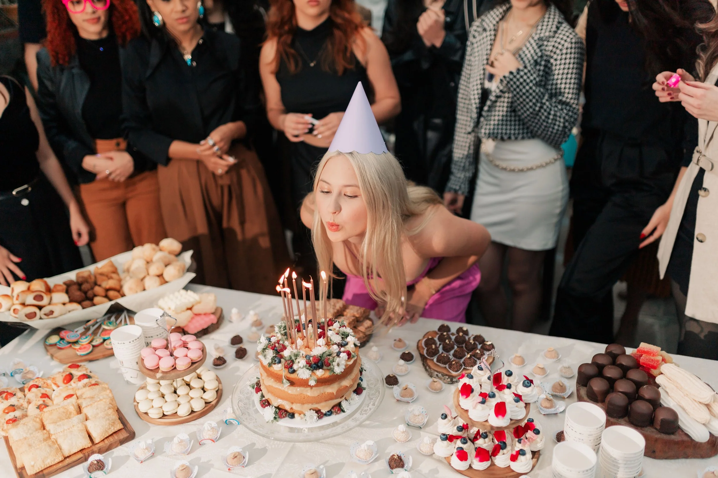 A woman wearing a blue party hat blows out candles on a decorated birthday cake at a celebration with a crowd gathered around a table of assorted desserts.