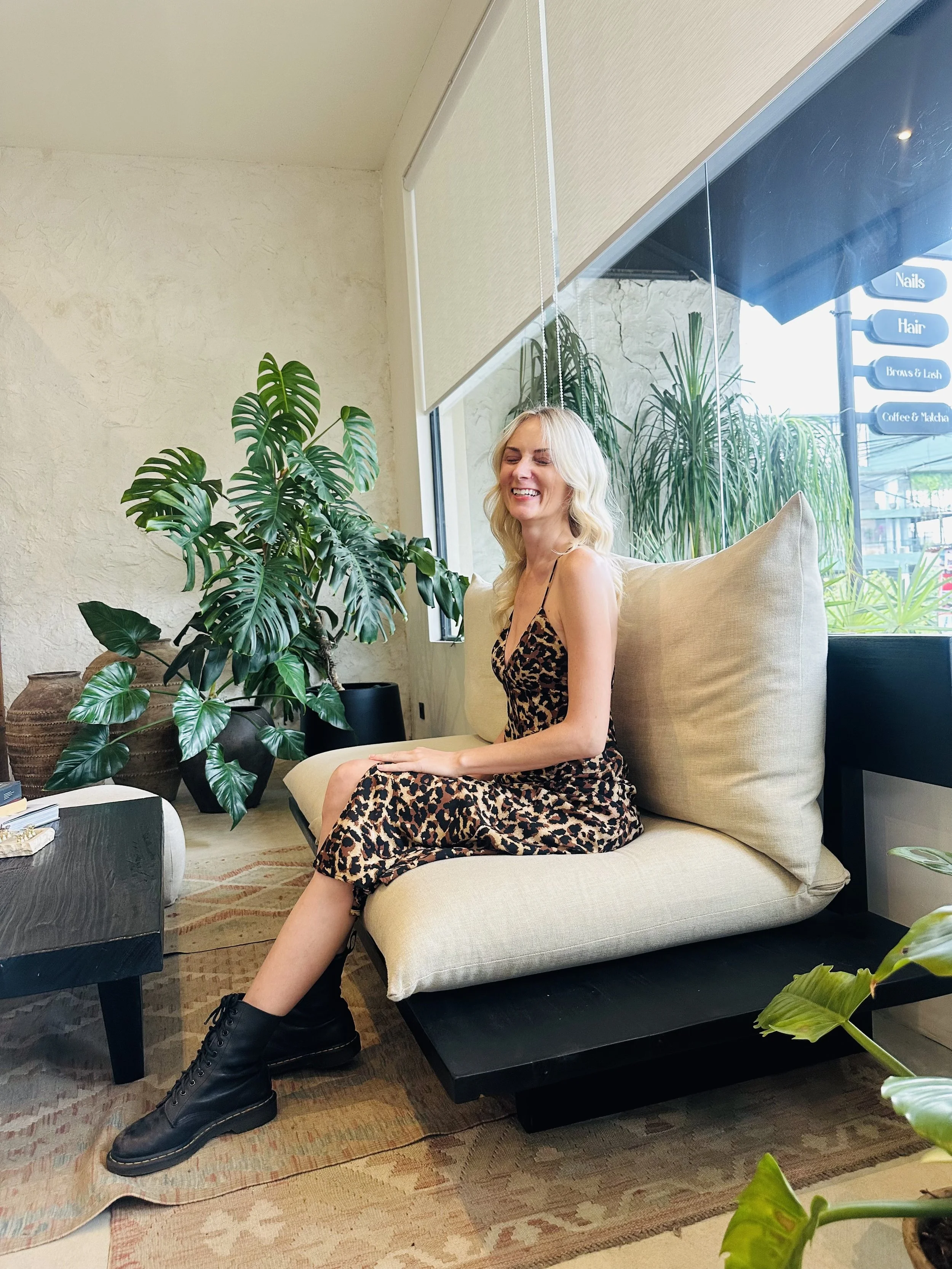 A young woman with blond hair wearing a leopard print dress, black boots, sitting on a beige sofa in a room with large green plants, a coffee table, and a window showing the street outside.