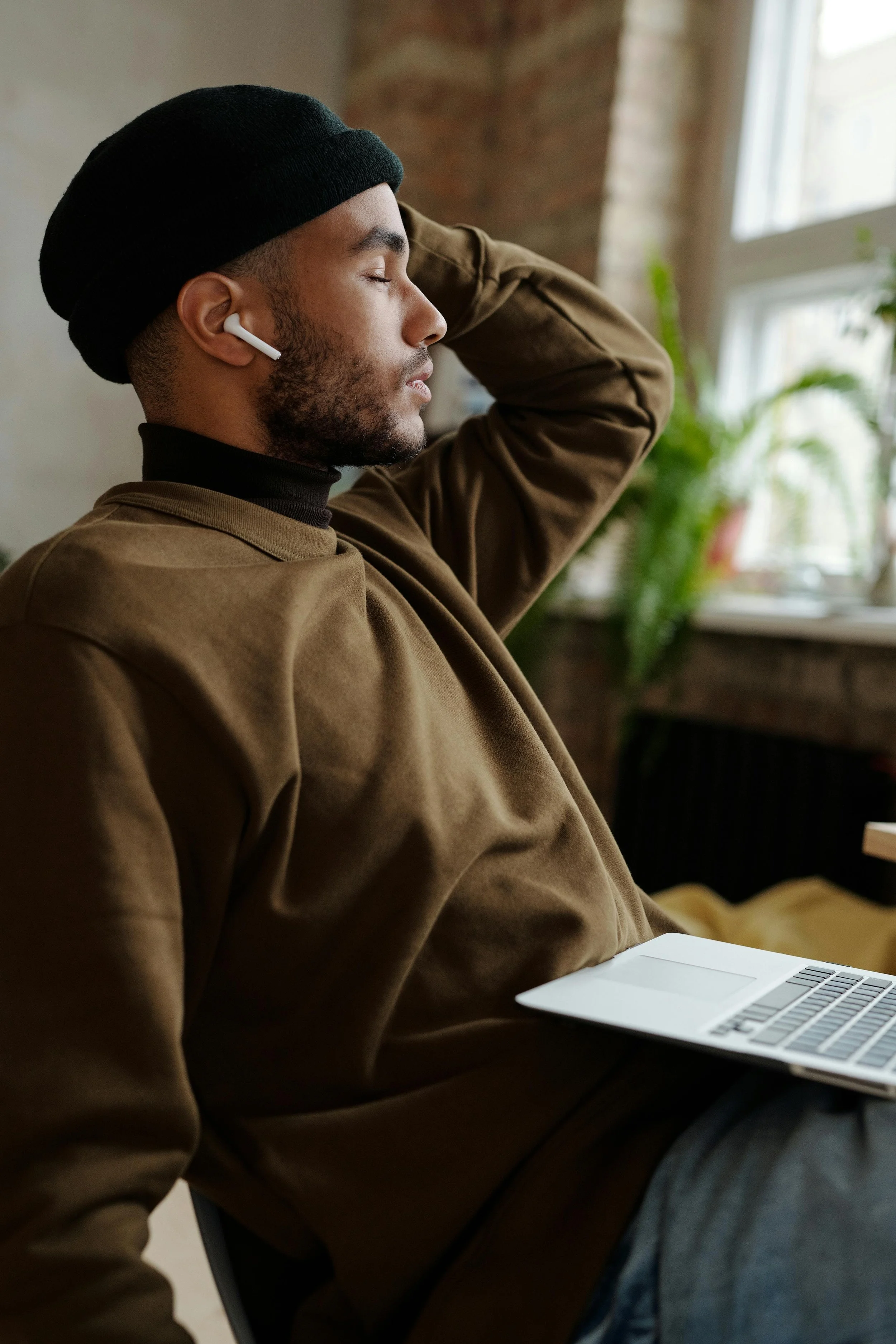 A man with a black beanie, earbuds, and a brown jacket, resting with his eyes closed, sitting at a table with a laptop in a room with a window and green plant in the background.