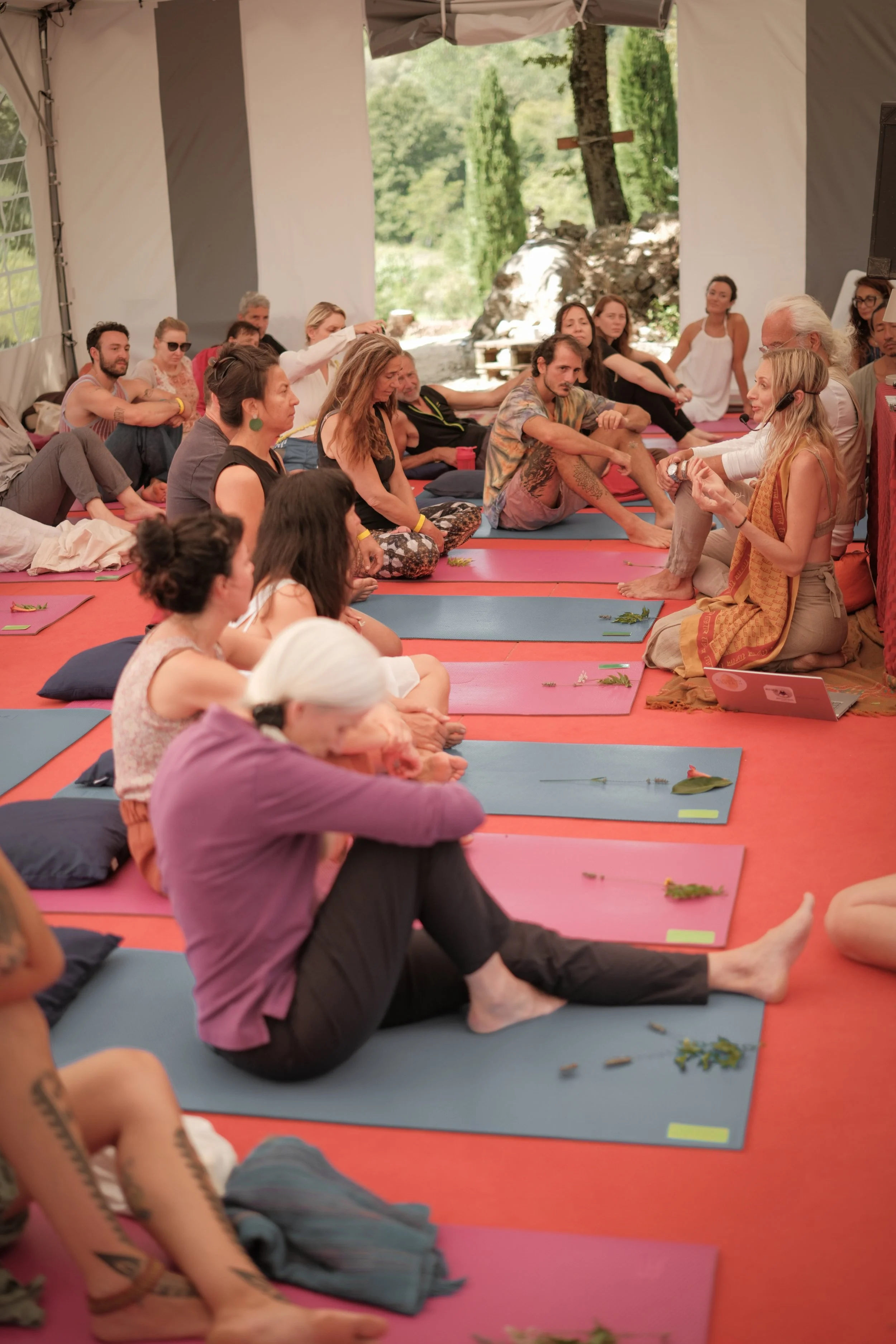 Group of people participating in a yoga or meditation session indoors, sitting on mats with leaves and small objects on the mats, in a tent with open sides overlooking a forested area.