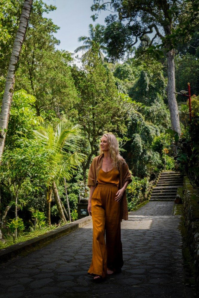 A woman walking on a stone path in a lush, green forest with tall trees and dense foliage.