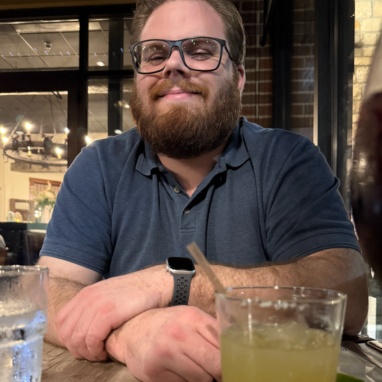 A smiling man with glasses and a beard sitting at a restaurant table with drinks, wearing a blue polo shirt and a smartwatch, in a cozy, warmly lit restaurant with large windows and exposed brick wall.