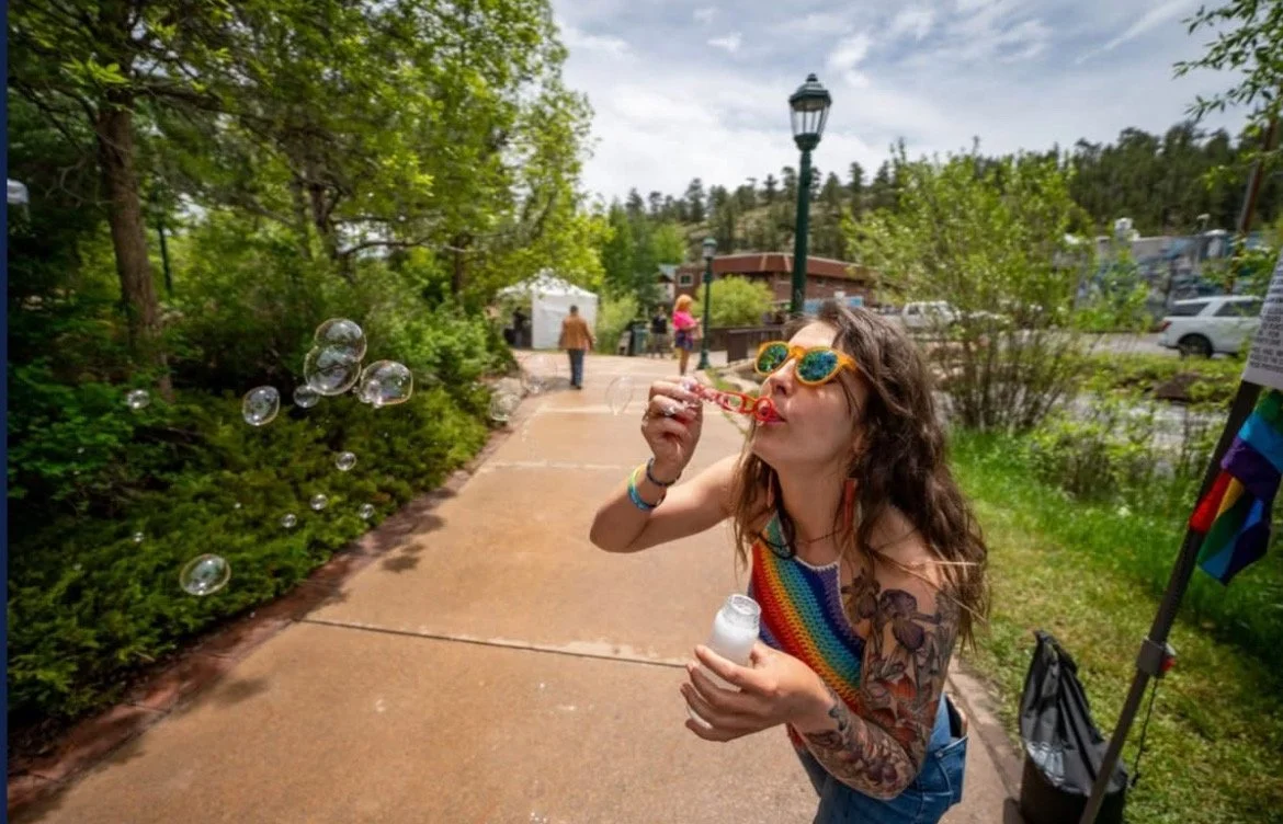 Woman with tattoos wearing rainbow-colored top and yellow sunglasses blowing bubbles on a walkway in a park.