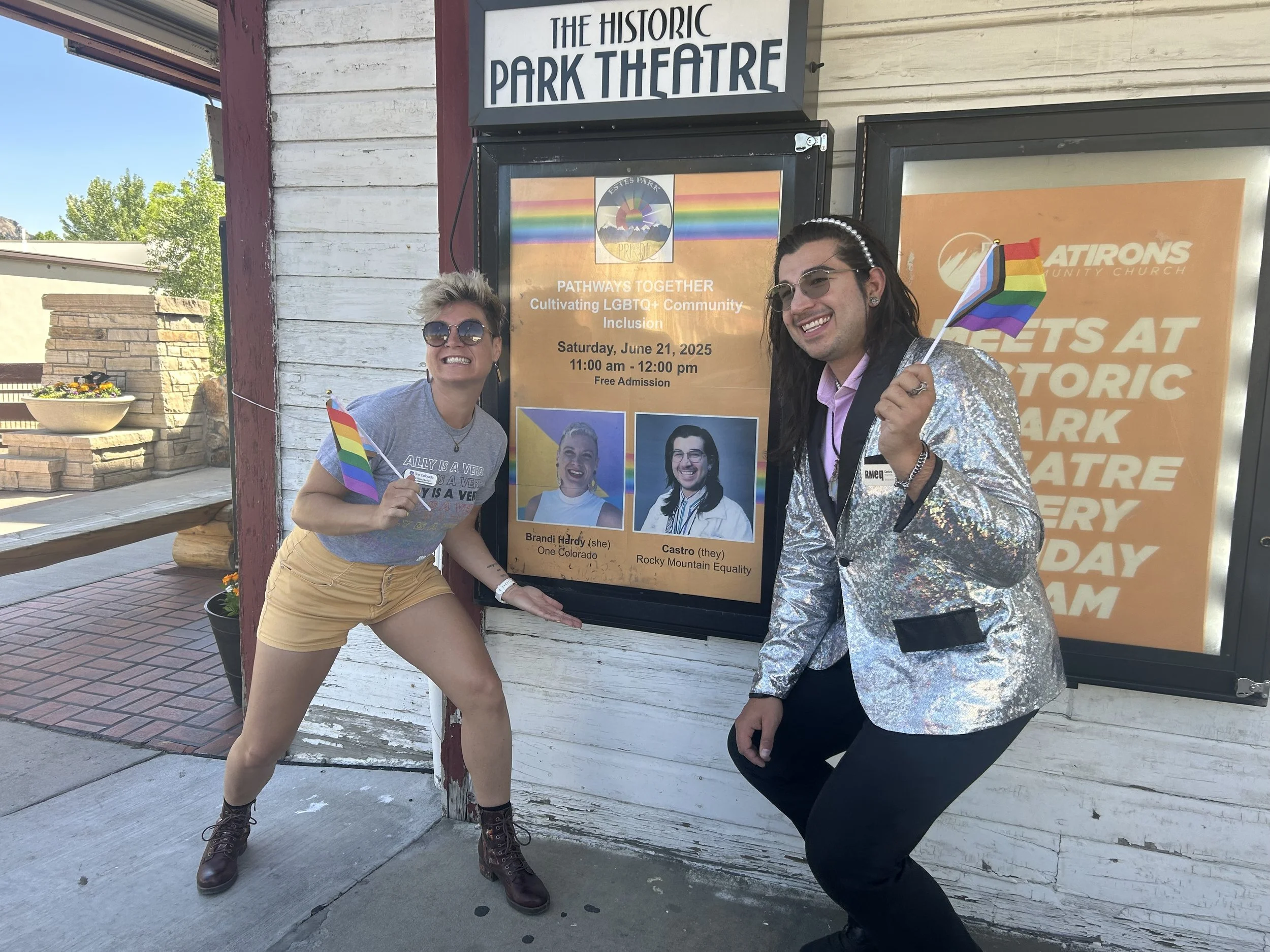 Two people smiling and posing in front of a sign for the historic Park Theatre, with small rainbow pride flags in their hands. One person is wearing sunglasses, a grey t-shirt, and yellow shorts, and the other is wearing glasses, a shiny silver jacket, and a pink shirt.