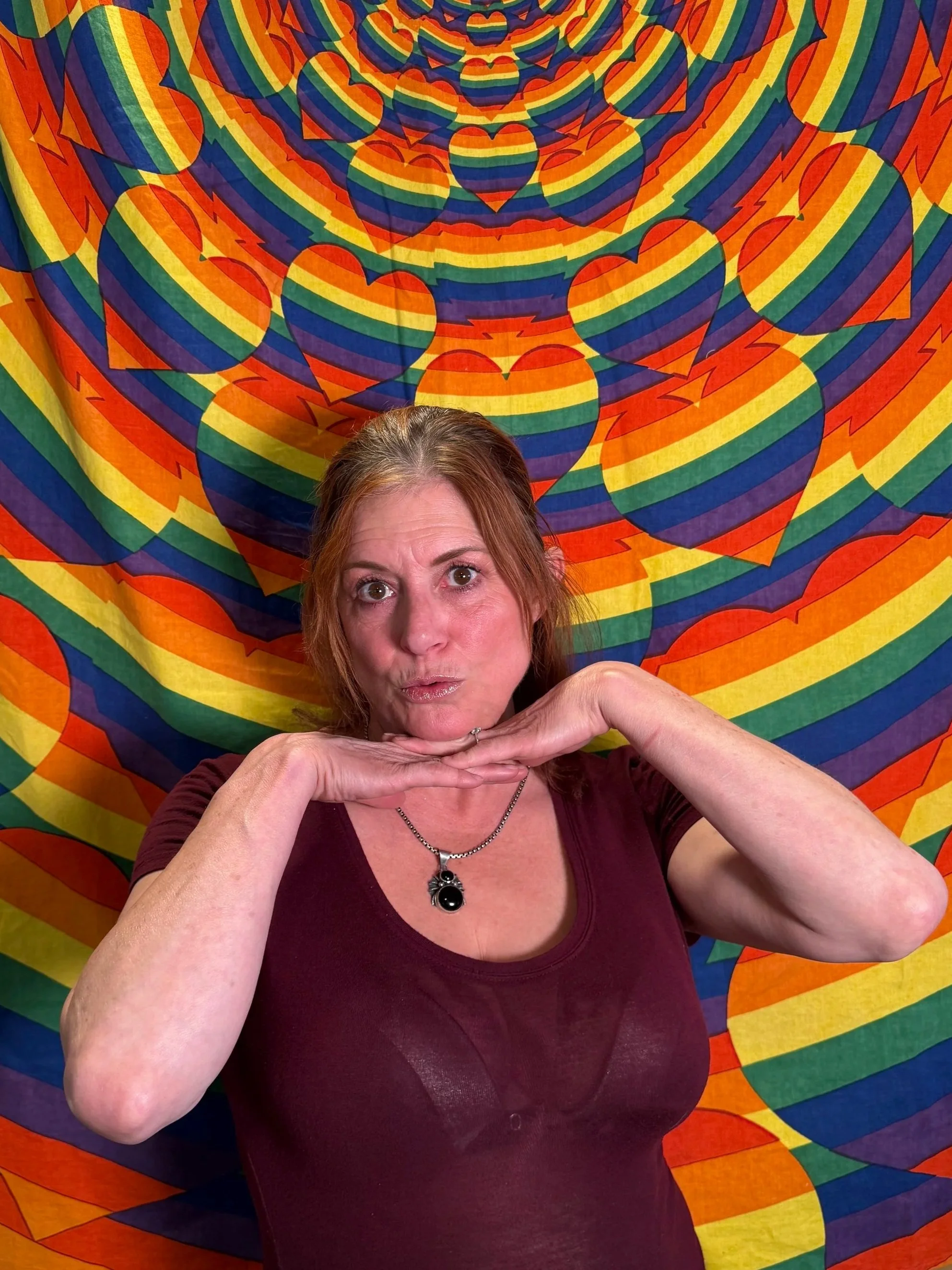 A woman with reddish hair posing with her chin resting on her hands in the foreground, with a colorful, psychedelic rainbow-colored heart and spiral pattern tapestry in the background.