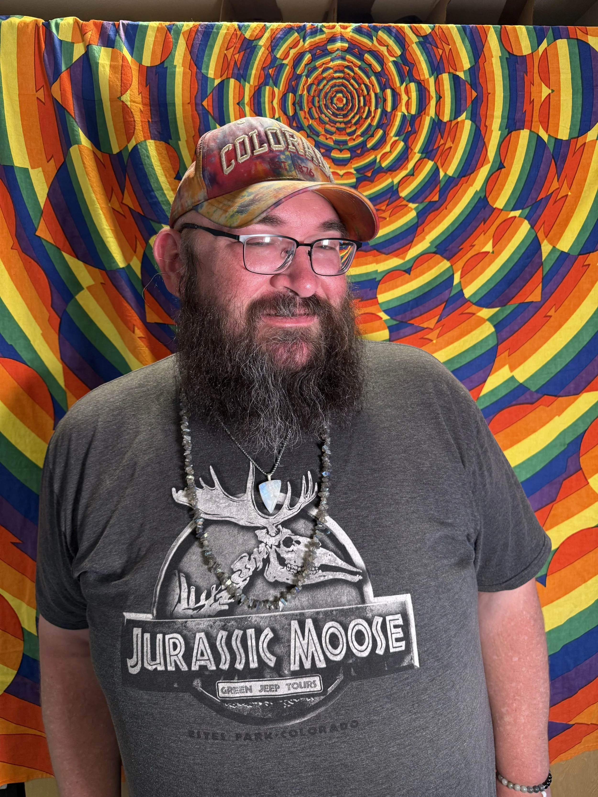 A man with glasses, wearing a colorful tie-dye cap with 'Colorado' written on it, and a black 'Jurassic Moose' T-shirt, standing in front of a psychedelic rainbow-colored tapestry.
