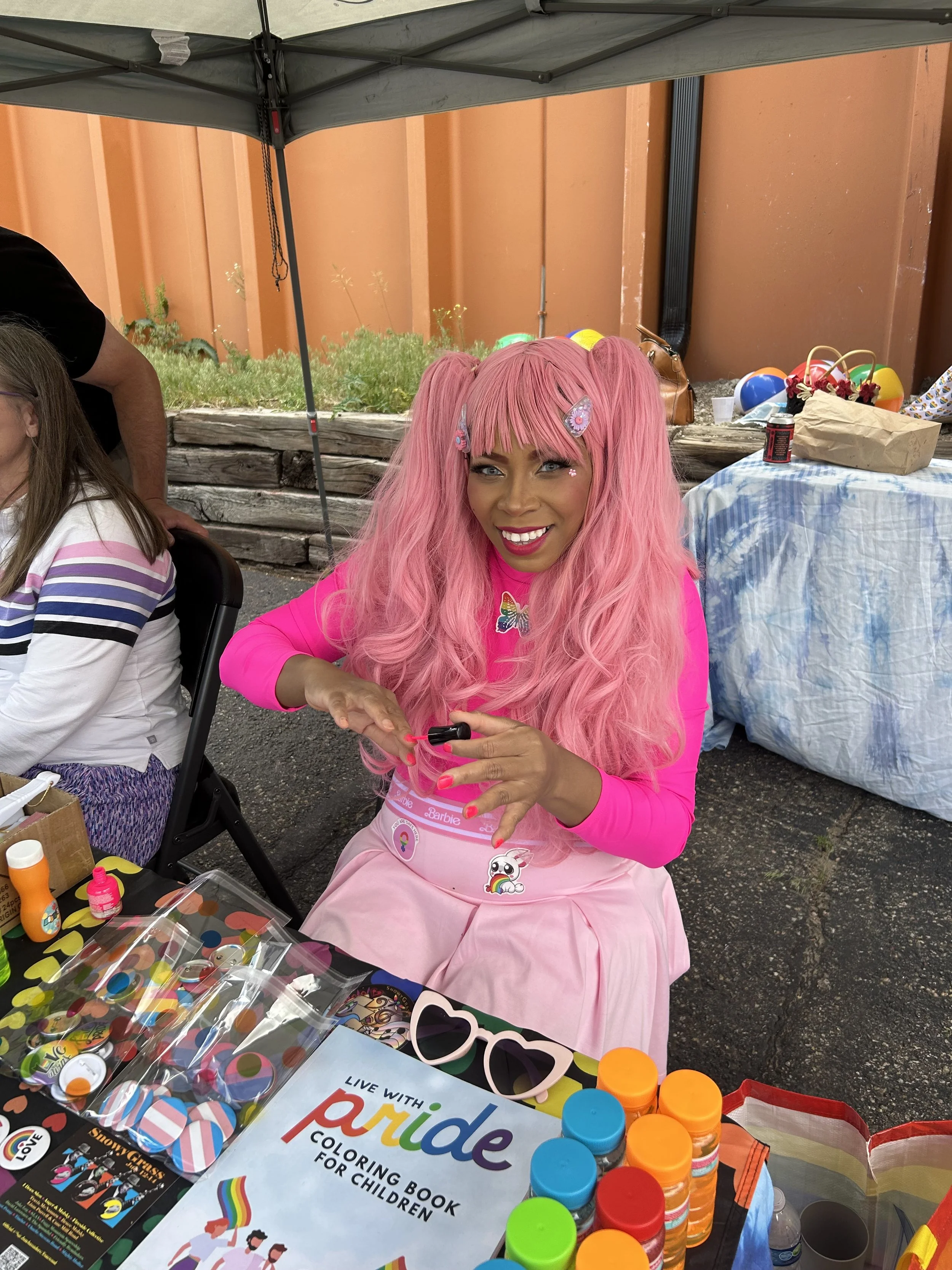 Woman with pink hair wearing pink apron with rainbow and unicorn stickers, sitting at a table with coloring art supplies and a coloring book for children, outdoors under a canopy.