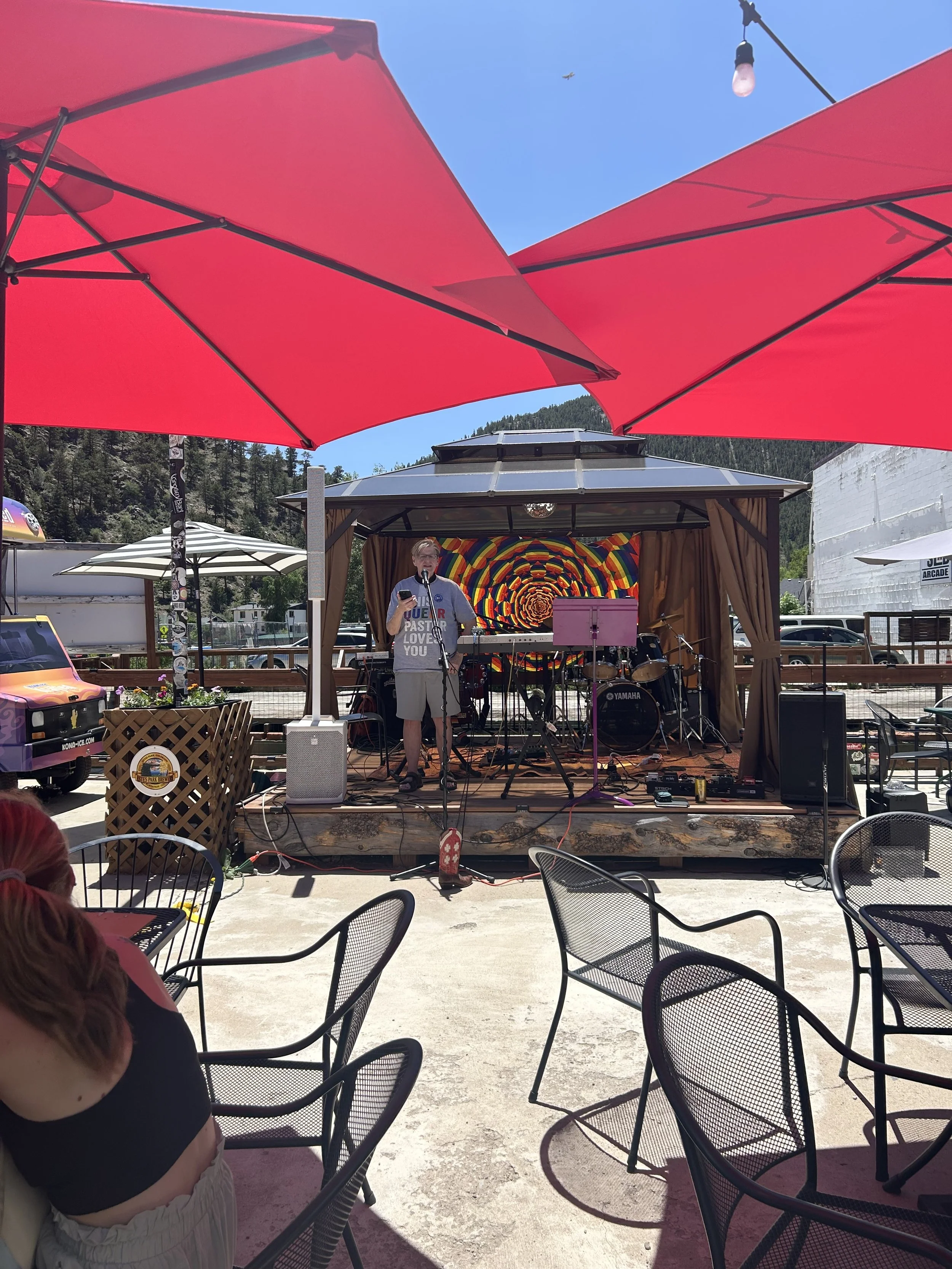 An outdoor music performance stage with a person singing into a microphone, surrounded by musical instruments, under a canopy with vibrant rainbow art in the background. Patio tables and chairs are in the foreground, with large red umbrellas providing shade.