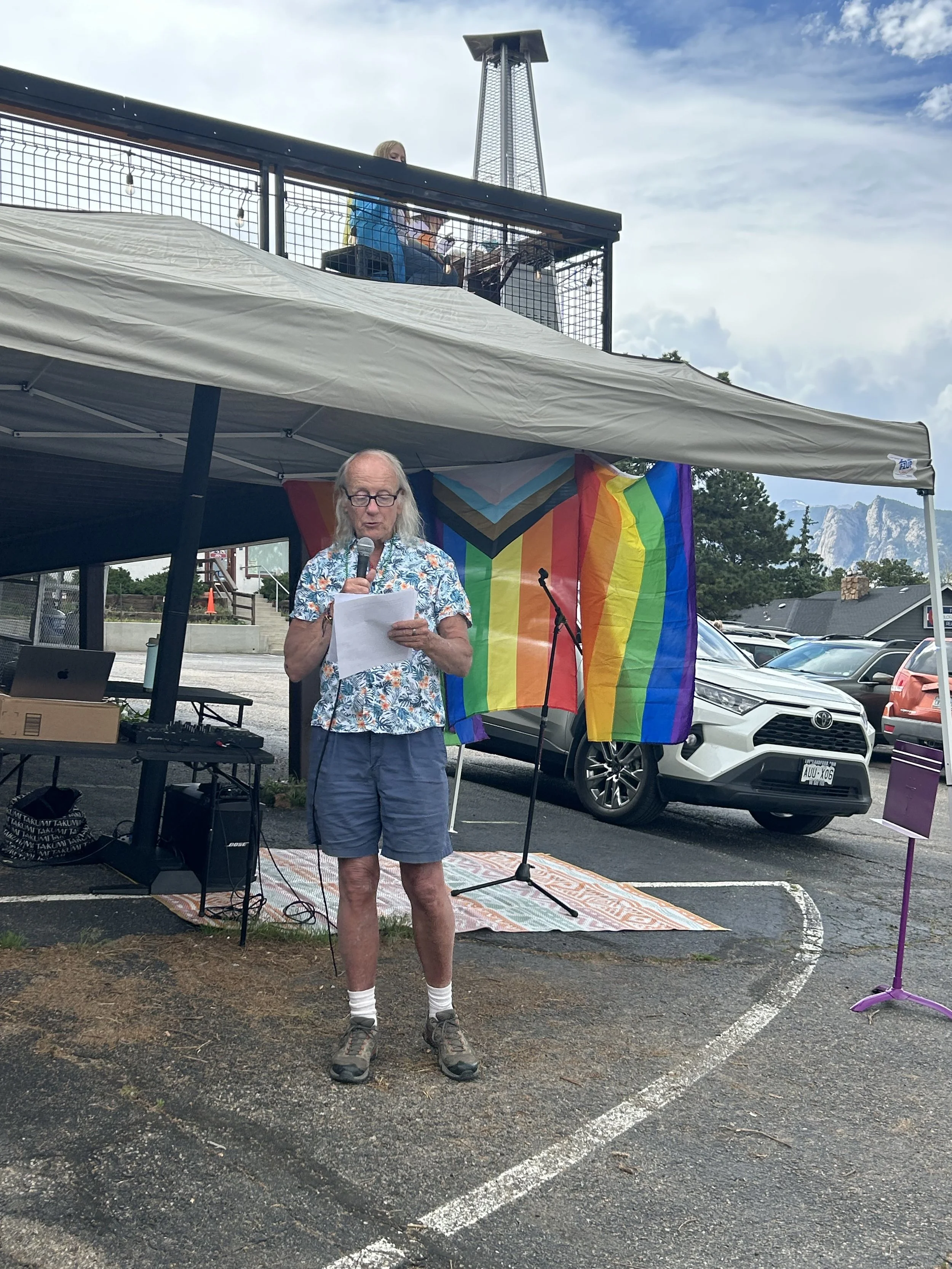 A person with long gray hair and glasses reading from a paper while holding a microphone during an outdoor event. Behind them, there is a rainbow pride flag hanging on a wall. The event is set in a parking lot with parked cars, and a mountain range is visible in the background.
