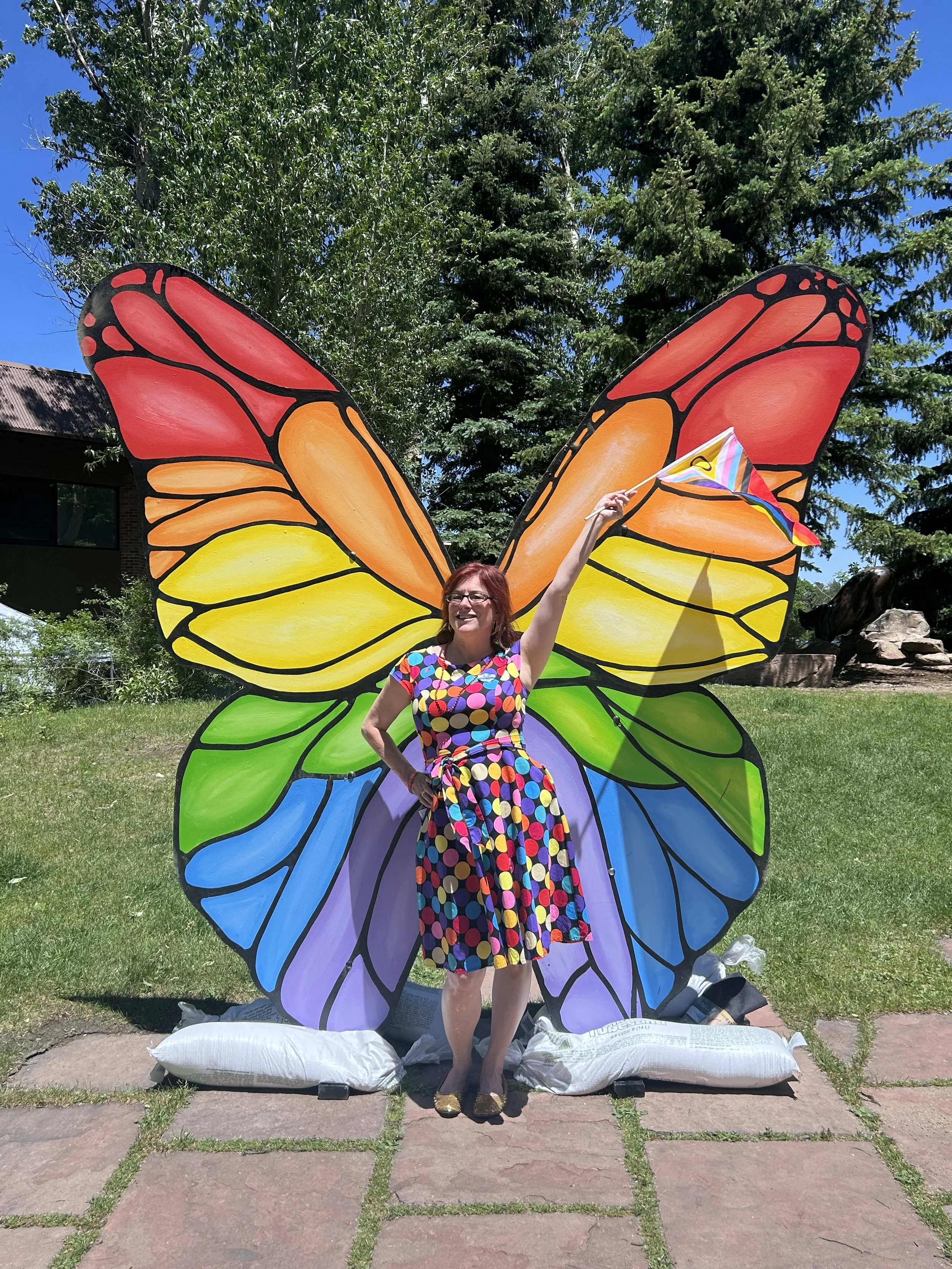 A woman standing in front of large, colorful butterfly wings outdoor during the daytime. She is holding a rainbow-colored flag and wearing a polka dot dress. The wings have bright colors, including red, orange, yellow, green, blue, and purple, with black outlines. There are trees and a building in the background.