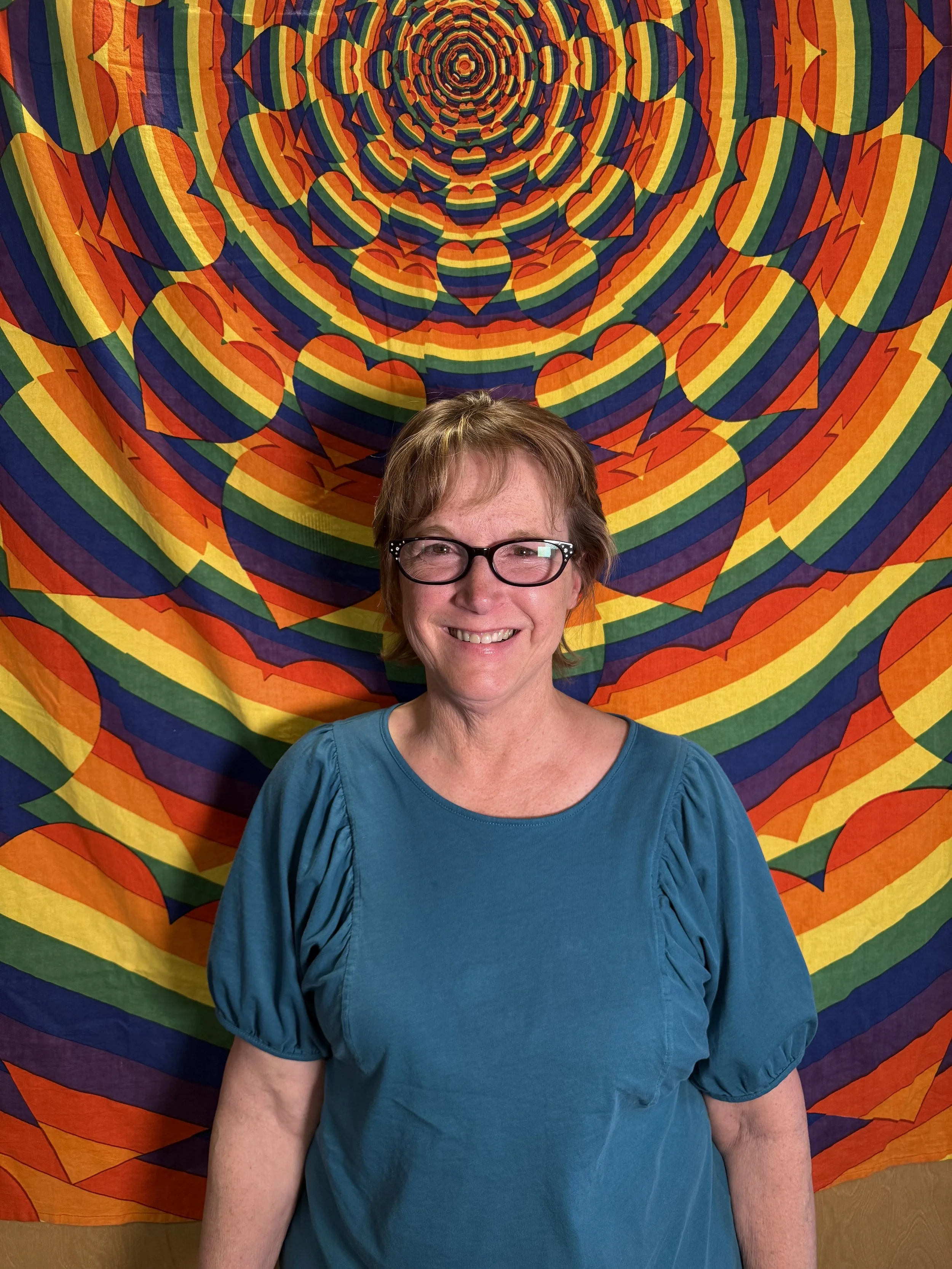 A woman with short brown hair and glasses smiling, standing in front of a colorful psychedelic tapestry with rainbow concentric heart and geometric patterns.