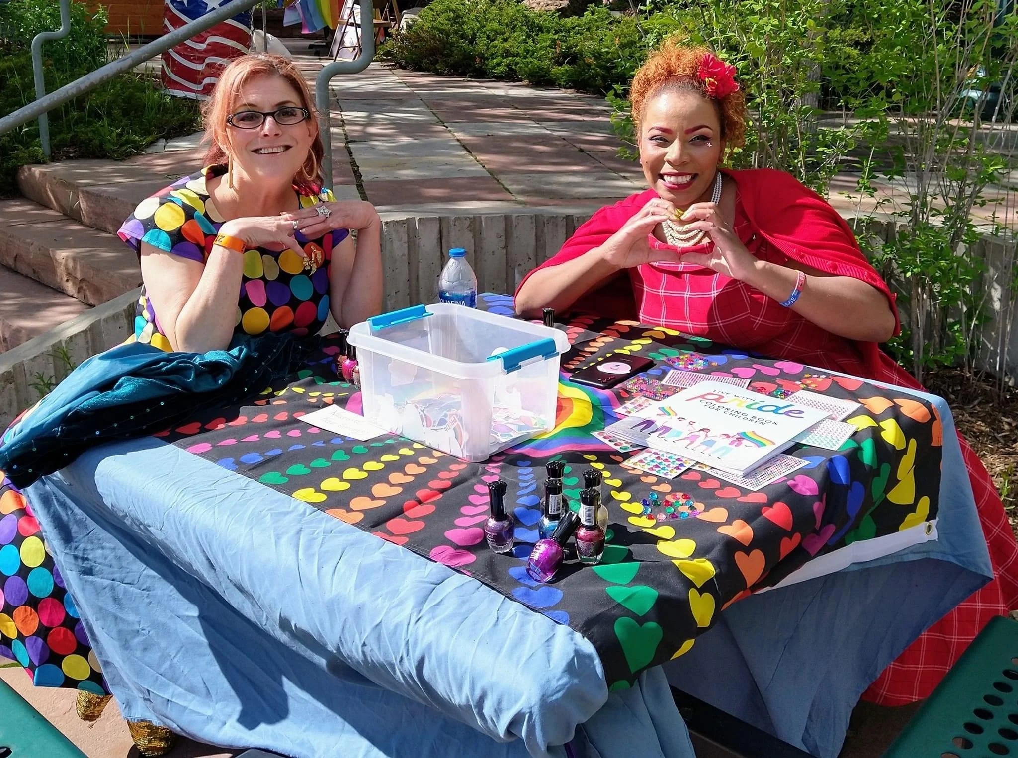 Two women smiling at a table with rainbow and heart-themed decorations, nail polish bottles, and informational flyers at an outdoor event.