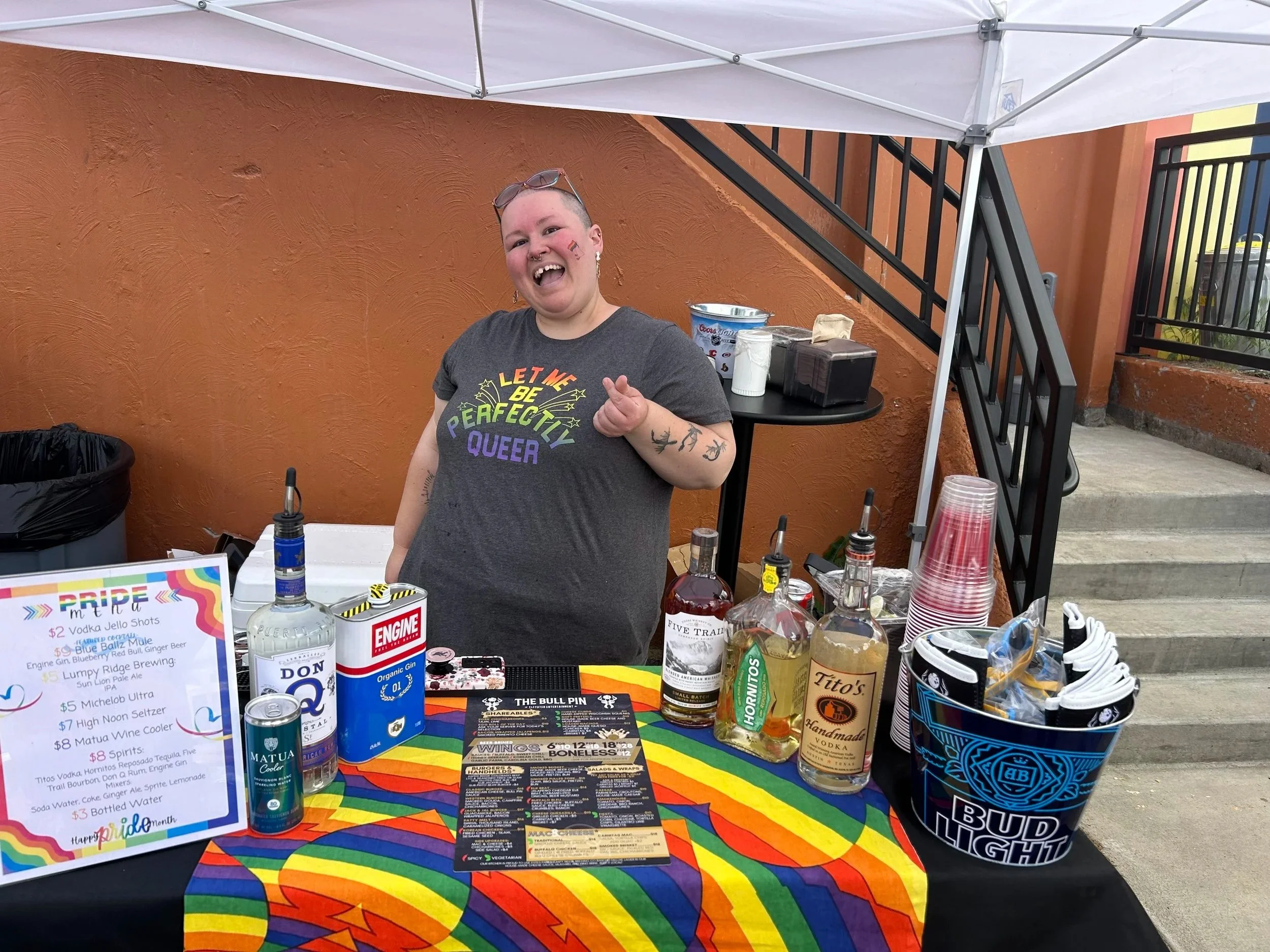A person with shaved head and tattoos on arm standing behind a colorful table at an outdoor event, smiling and wearing a gray T-shirt with rainbow-colored text.