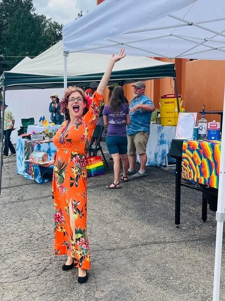A woman in an orange floral dress smiling and waving at an outdoor event, standing on cracked pavement near a white tent with tables and people in the background.
