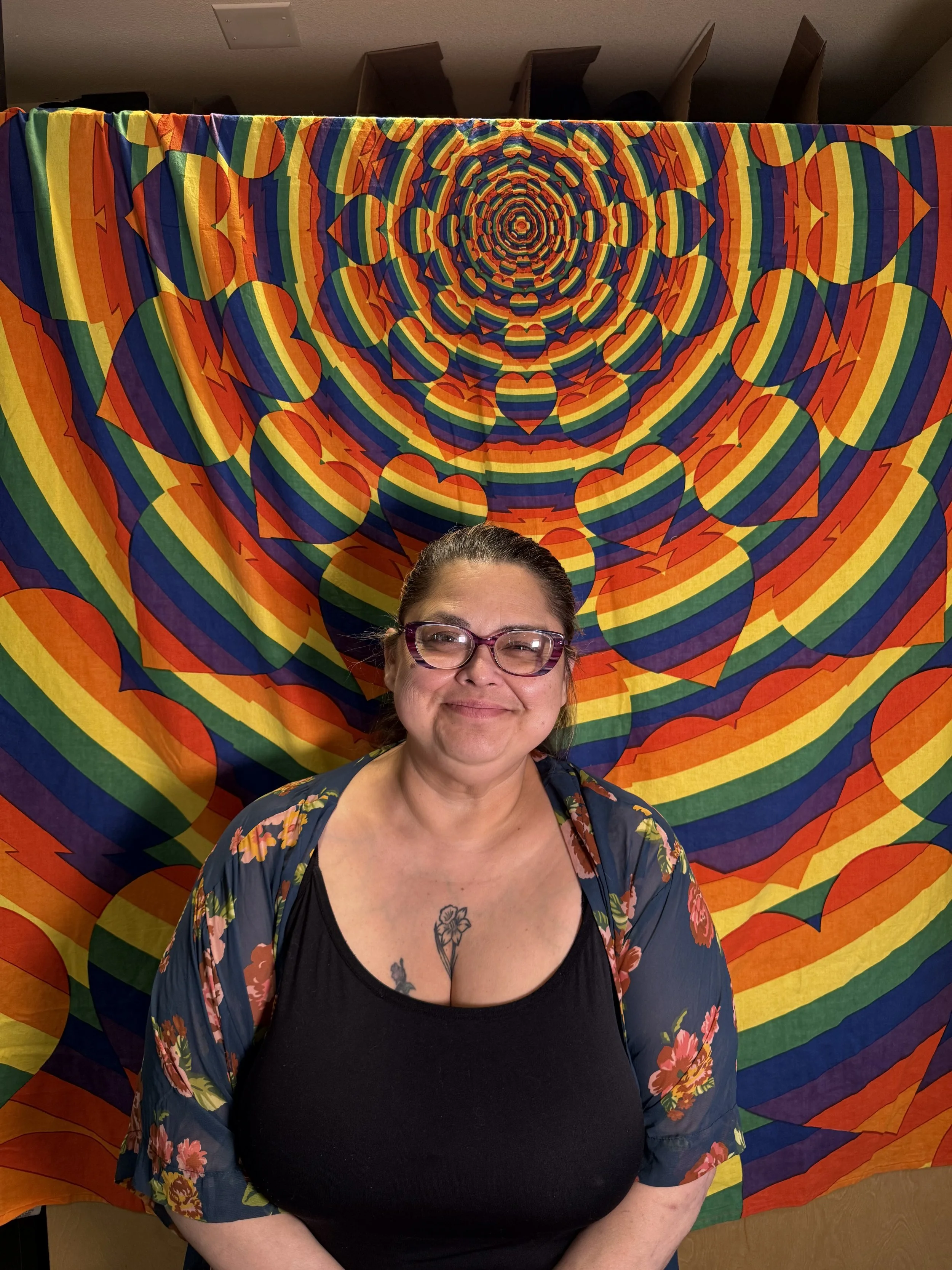 A woman with dark hair wearing glasses and a floral shirt with a low neckline, smiling, standing in front of a colorful, hypnotic rainbow pattern tapestry hung on a wall.