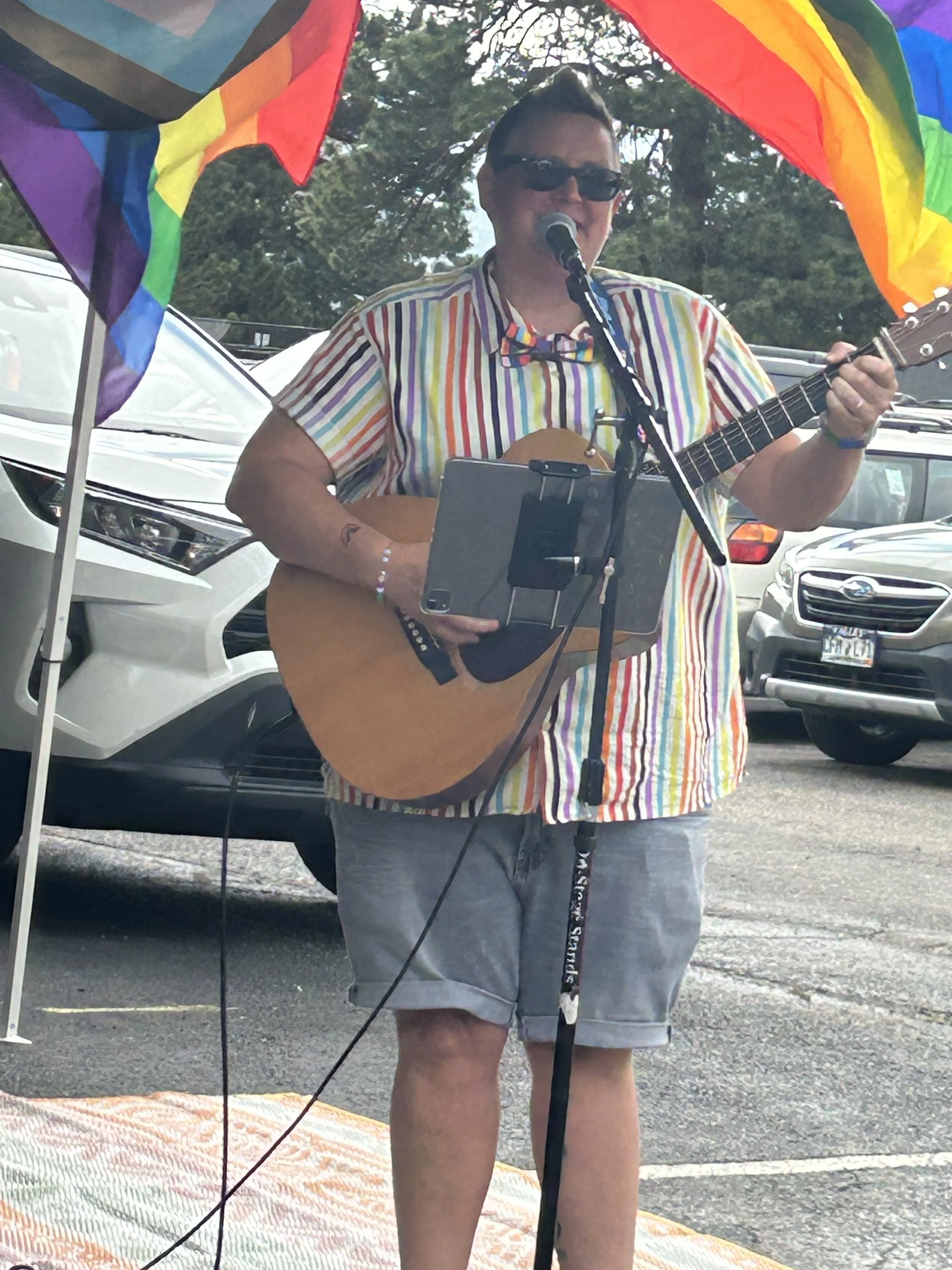 A man wearing sunglasses, a rainbow-striped shirt, and khaki shorts playing an acoustic guitar at an outdoor event, with a colorful rainbow flag and parked cars in the background.