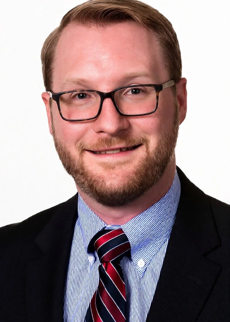 Headshot of a man with glasses, a beard, wearing a suit and striped tie, smiling.