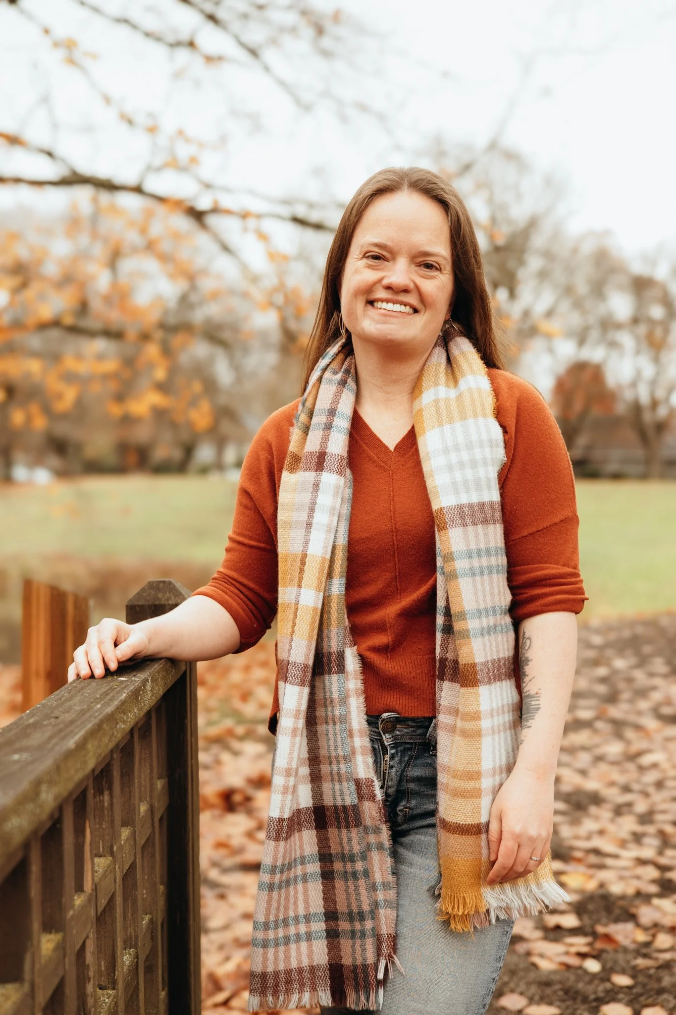 A woman smiling outdoors during fall, wearing an orange sweater, jeans, and a plaid scarf, standing by a wooden fence with fallen leaves on the ground and trees with autumn foliage in the background.