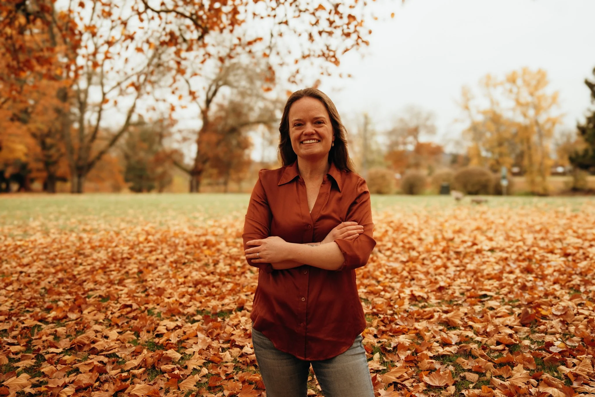 A woman standing in a park filled with fallen autumn leaves, smiling with arms crossed, surrounded by trees with orange, yellow, and brown leaves.