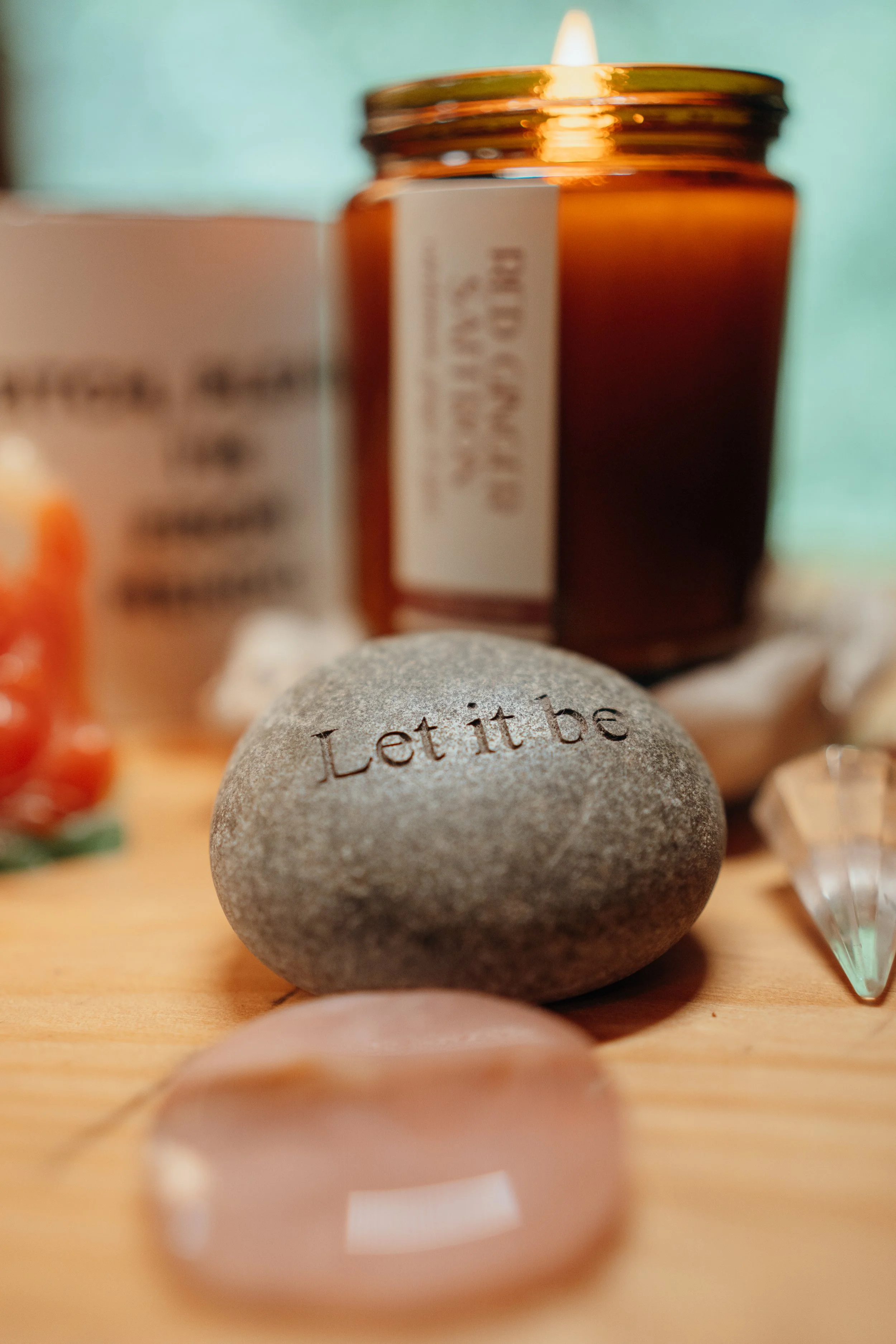 Close-up of a gray stone with the words "Let it be" engraved on it. In the background, there are blurred objects including a brown glass jar, a candle, and other items on a wooden surface.