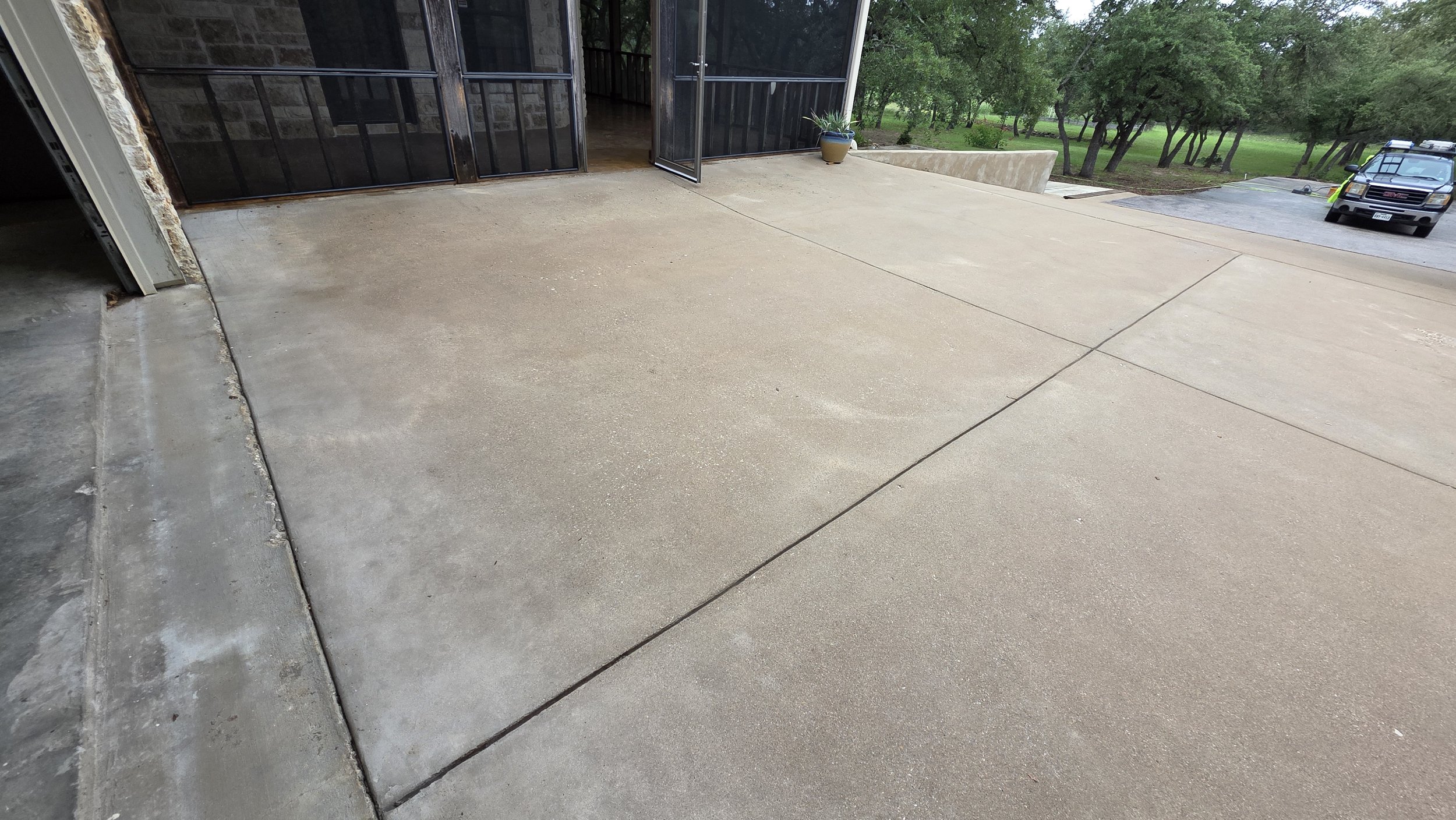 A concrete driveway with a turning area, leading to a house with a sliding glass door and a potted plant near the entrance. There is a parked black pickup truck and some trees in the background.