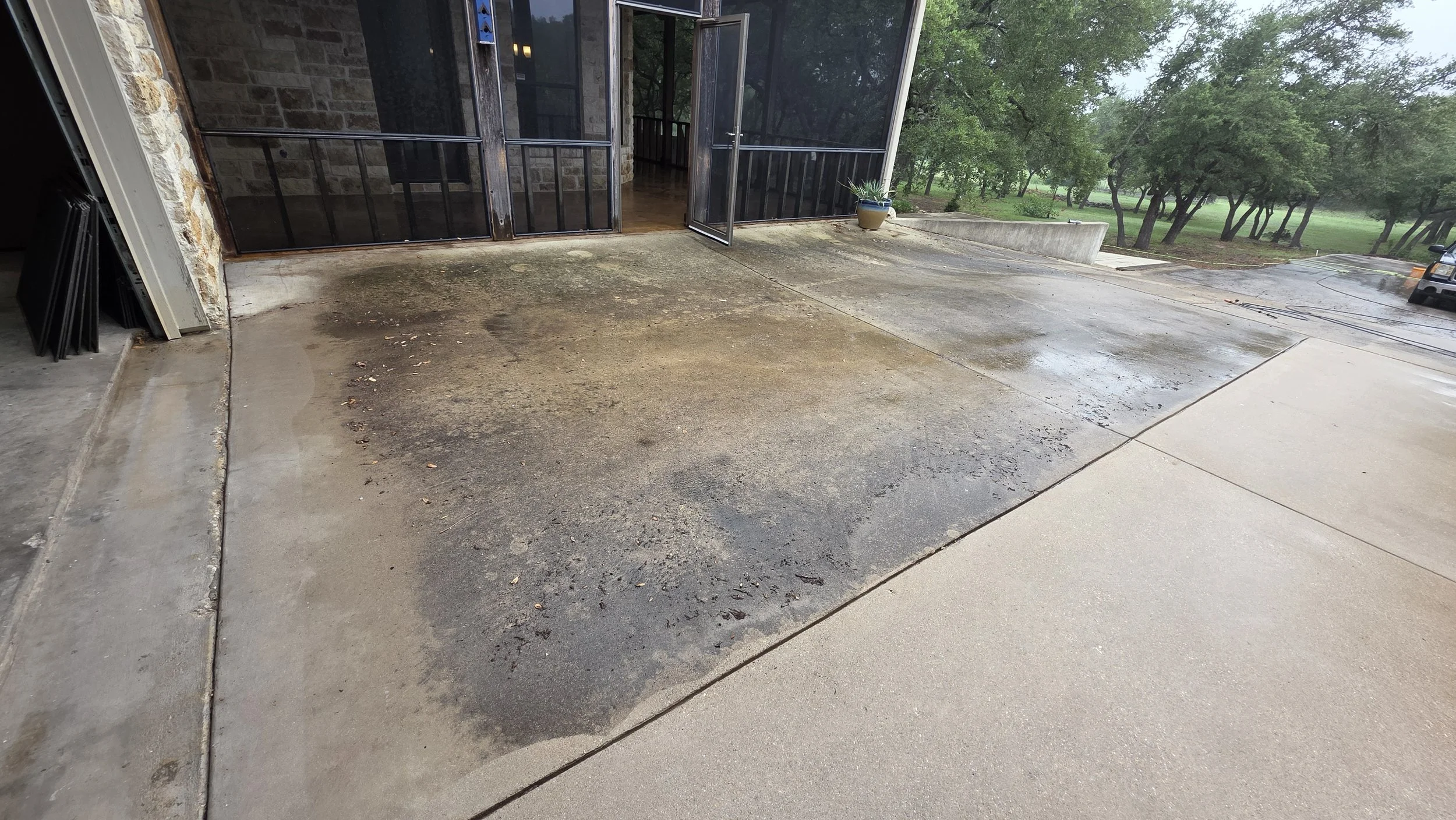 Wet and dry concrete patio area outside with a potted plant, open screened door leading inside, and a grassy park with trees in the background.