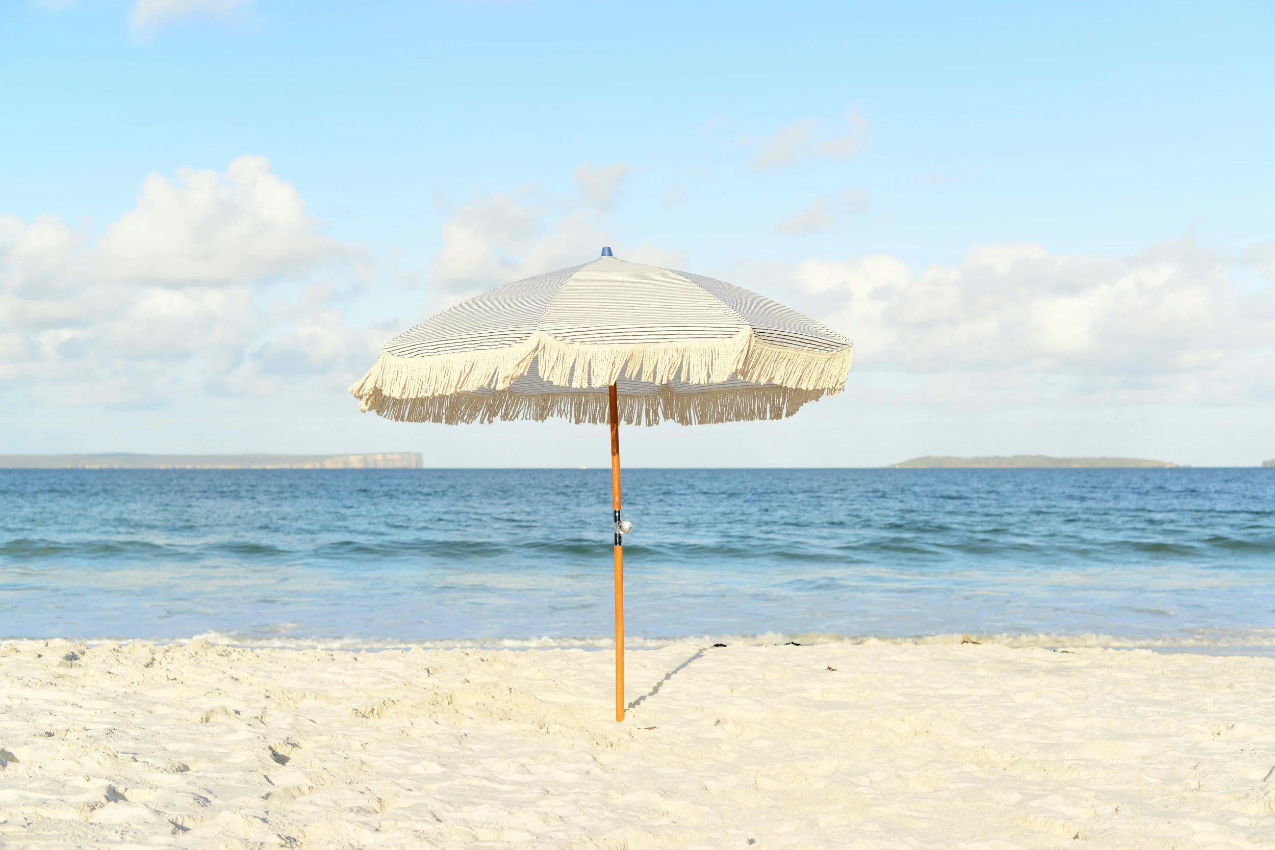 A beach scene with white sand, a single beige beach umbrella, ocean water in the background, and a partly cloudy sky.