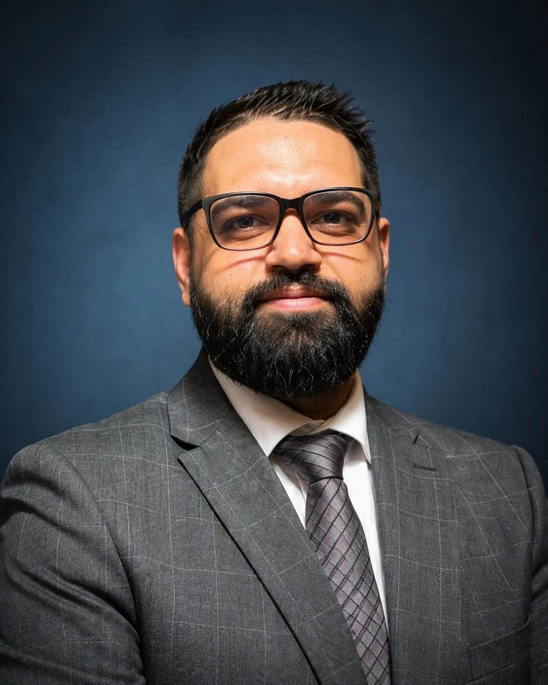 Professional man with glasses, beard, and dark hair, wearing a gray suit with a checkered pattern and a matching tie, posing against a dark blue background.