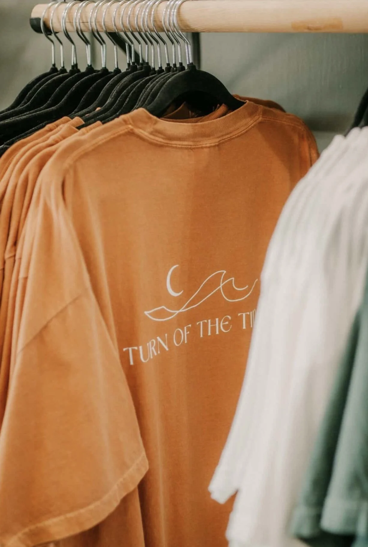 Orange T-shirt hanging on a black hanger with white text and a graphic, among other black and white T-shirts on a clothing rack.