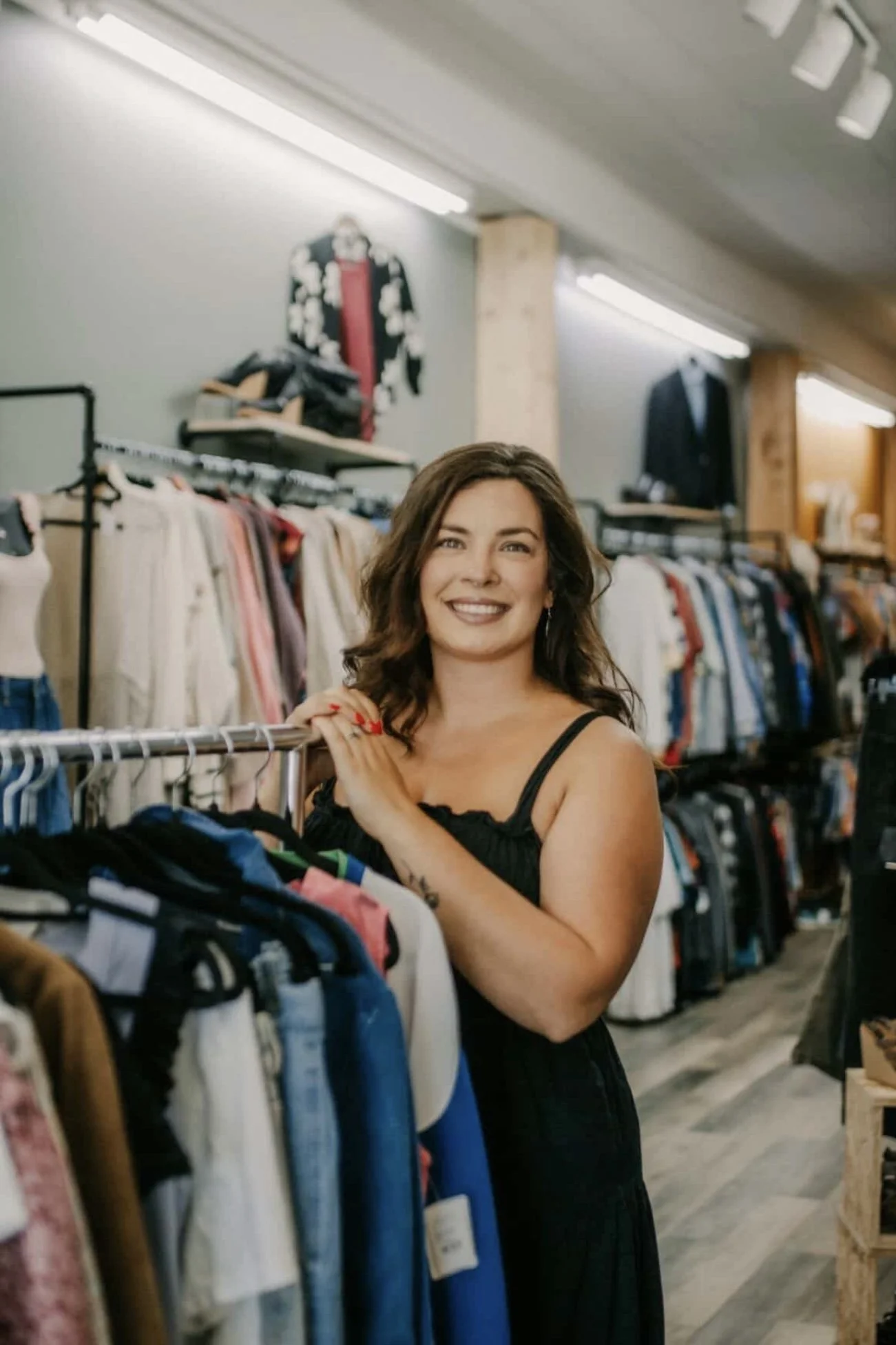 A smiling woman with wavy brown hair and a black sleeveless top shopping in a clothing store with racks of various clothes around her.