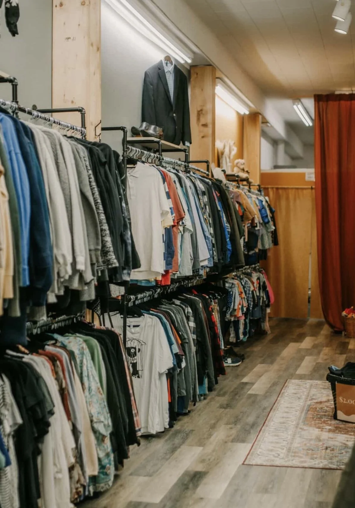 A clothing store with racks of various shirts and jackets, wooden walls, a dark-colored curtain, and a small rug on the floor.