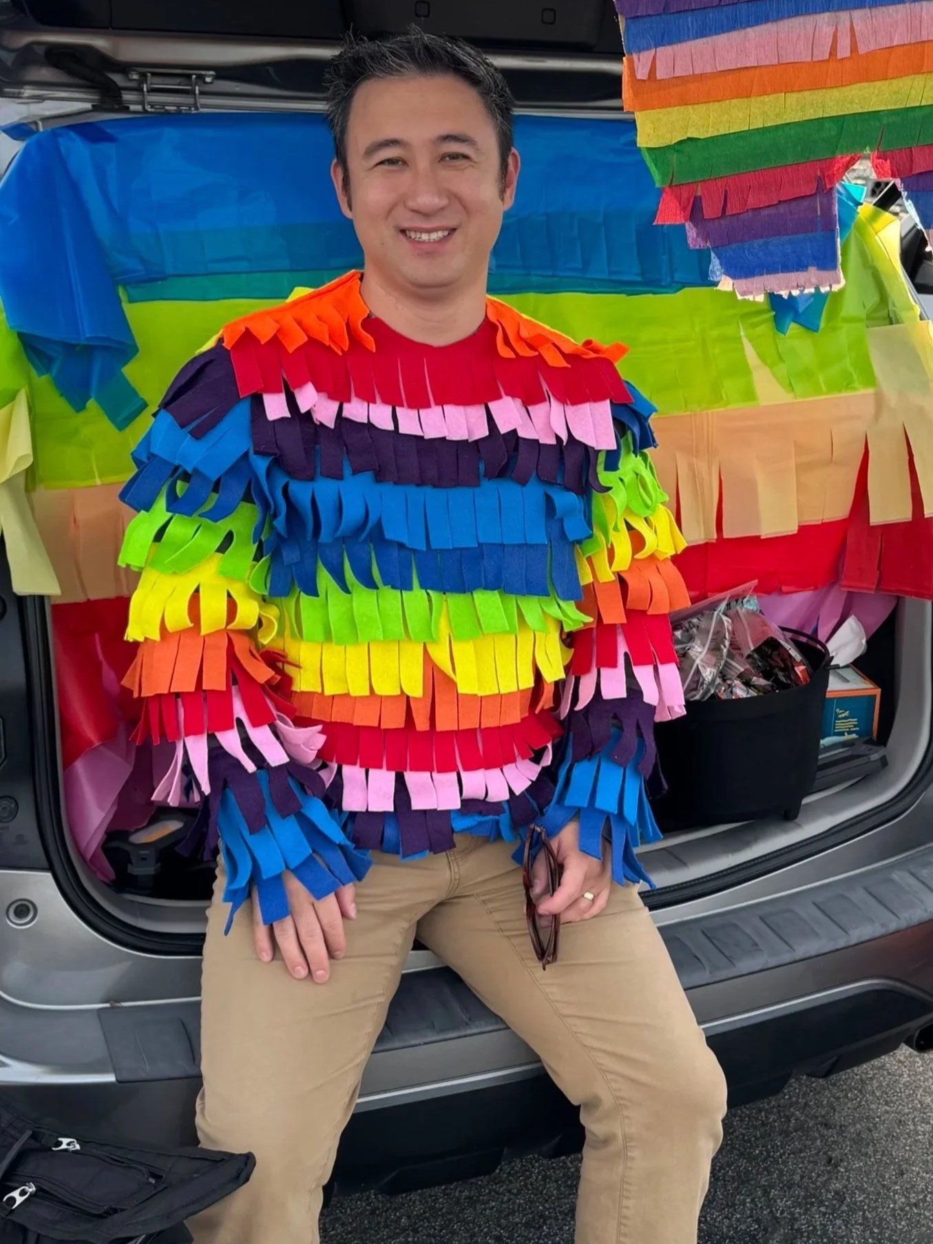 A man dressed in a rainbow-colored costume made of fringed paper strips stands in front of an open vehicle trunk decorated with rainbow-colored paper strips. The man is smiling, sitting with legs apart, holding glasses in his right hand and resting his left hand on his knee. The setup suggests a festive, colorful celebration, possibly related to Pride.