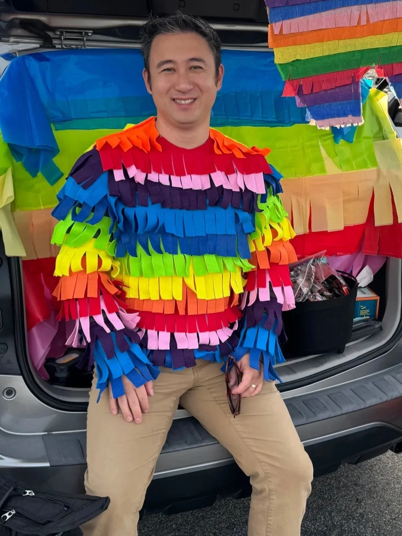 A man dressed in a rainbow-colored costume made of fringed paper strips stands in front of an open vehicle trunk decorated with rainbow-colored paper strips. The man is smiling, sitting with legs apart, holding glasses in his right hand and resting his left hand on his knee. The setup suggests a festive, colorful celebration, possibly related to Pride.